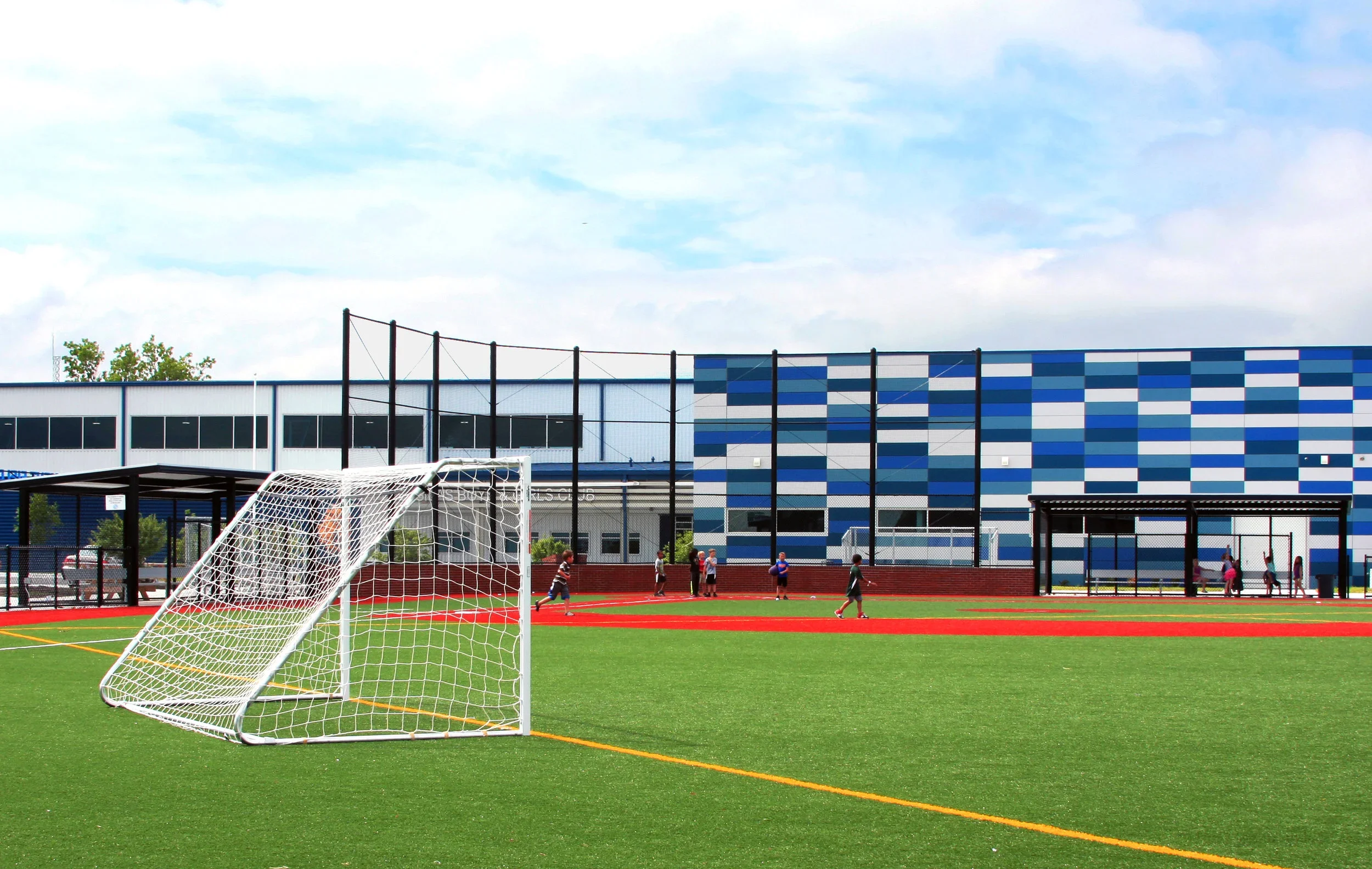 A soccer field with a goalpost in the foreground and children playing in the background, adjacent to a modern building with a blue tiled exterior.