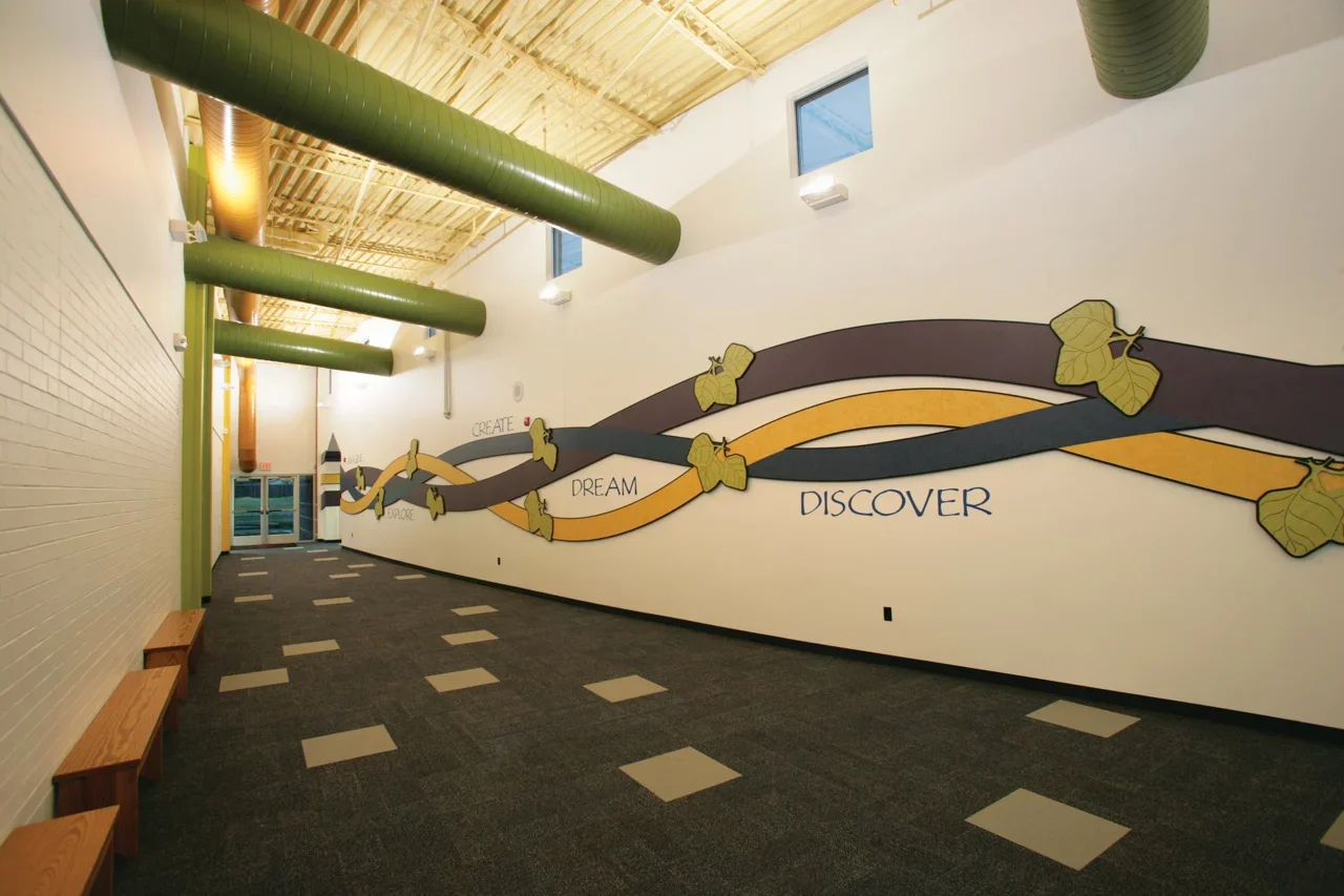 Interior hallway with green ductwork on the ceiling, a white brick wall on the left, and colorful mural artwork with words "Create," "Explore," "Dream," and "Discover" on the right wall, featuring a winding ribbon design with leaf embellishments.