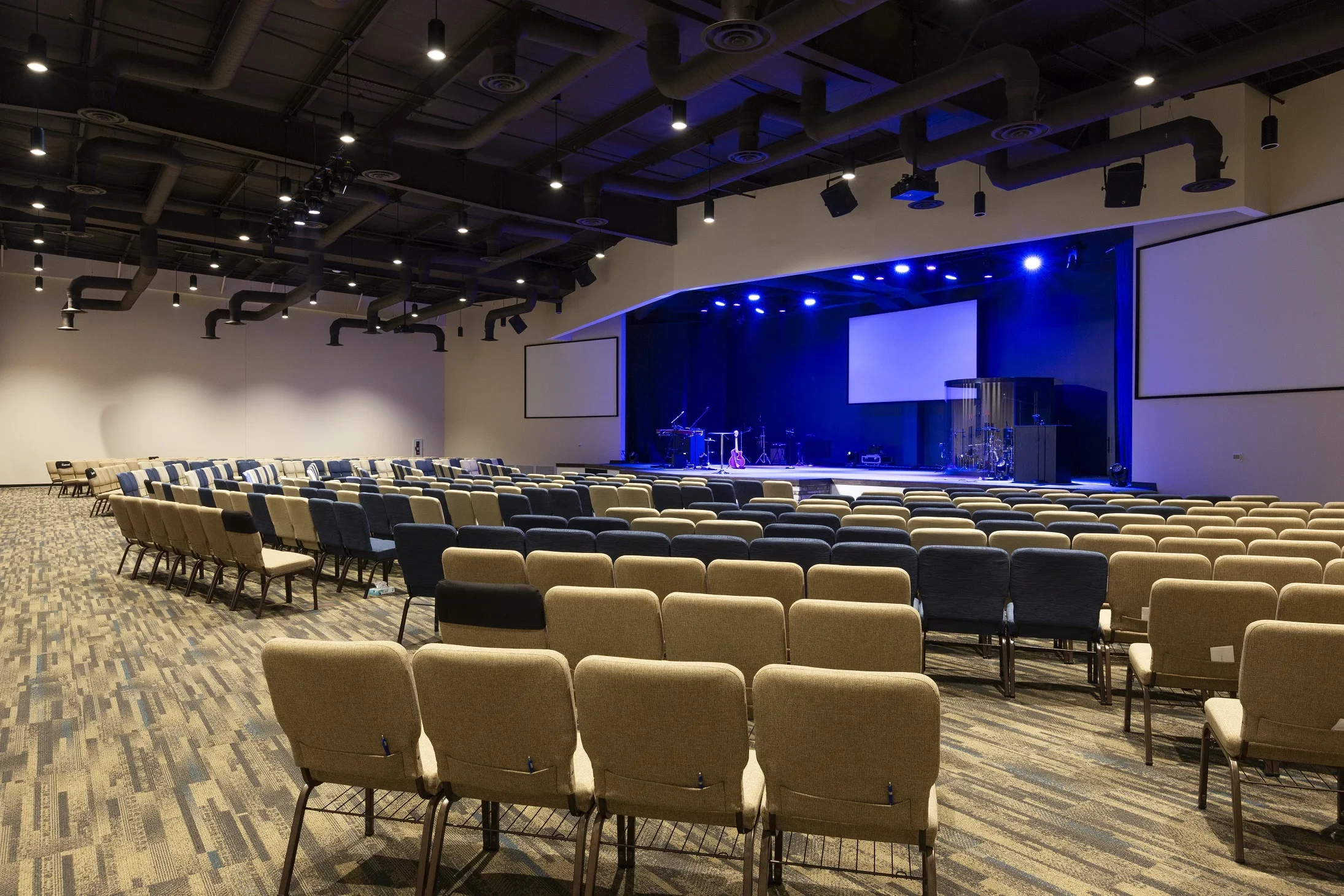 Empty auditorium with rows of beige and blue chairs facing a stage illuminated with blue lights, instruments on stage, and two large screens on either side.