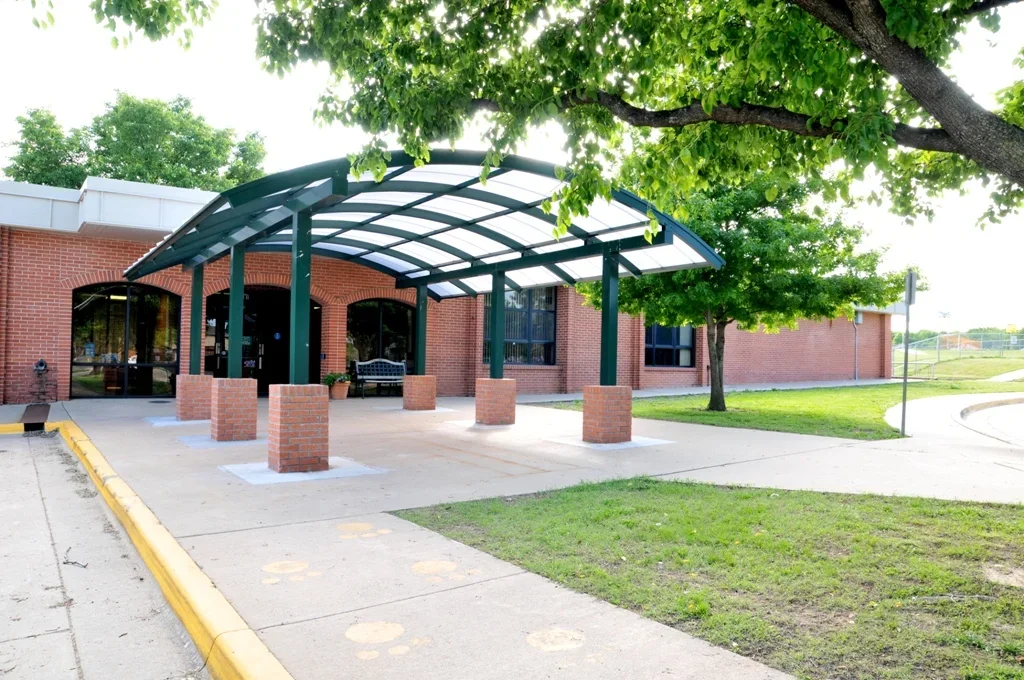 Exterior of Wayside Elementary in Bartlesville, OK with a covered entrance supported by green metal beams, surrounded by green trees and grass, with a sidewalk and parking lot nearby.