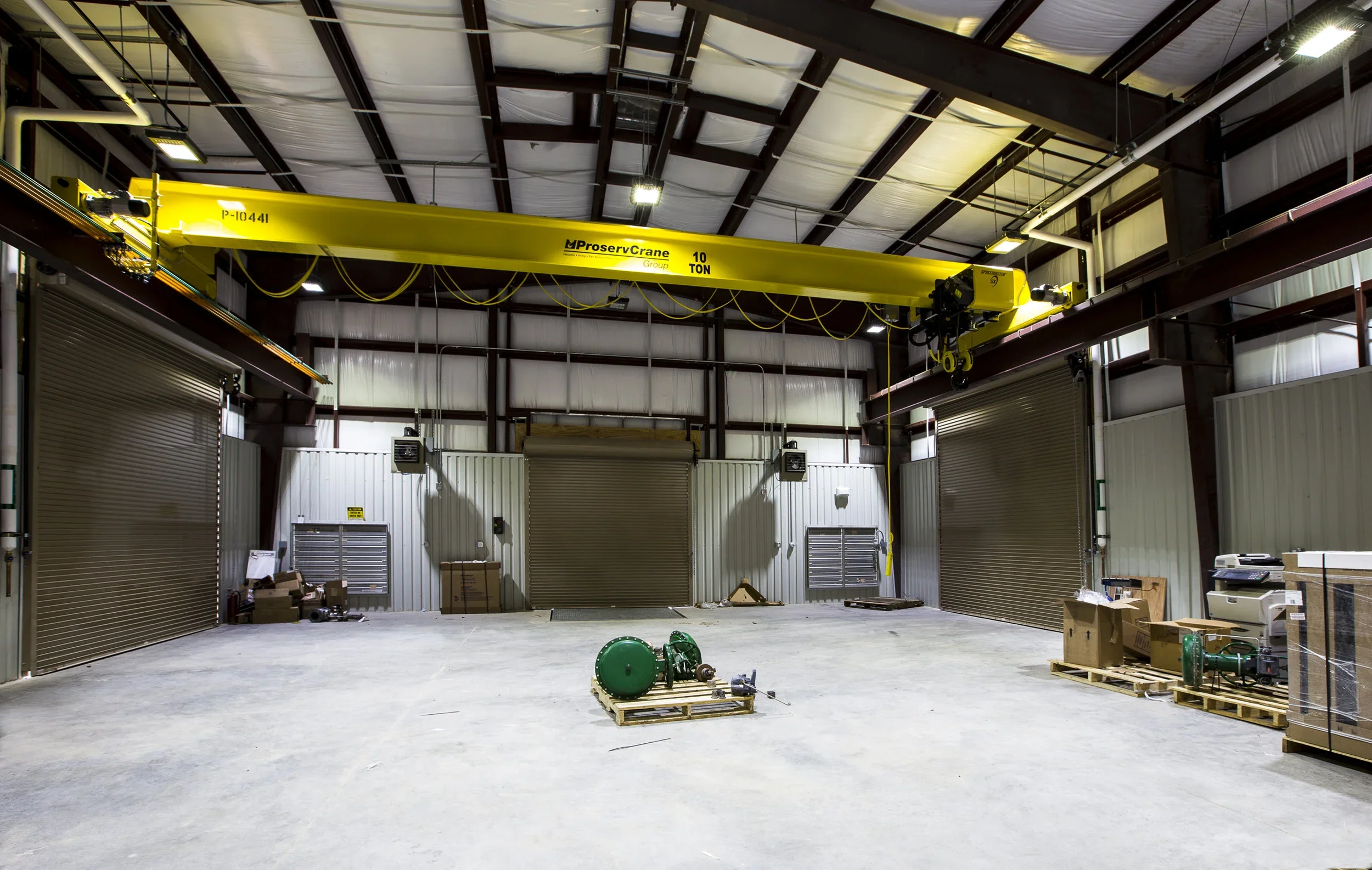 Empty industrial warehouse with five roll-up doors, a yellow overhead crane, and various boxes and equipment on the floor.