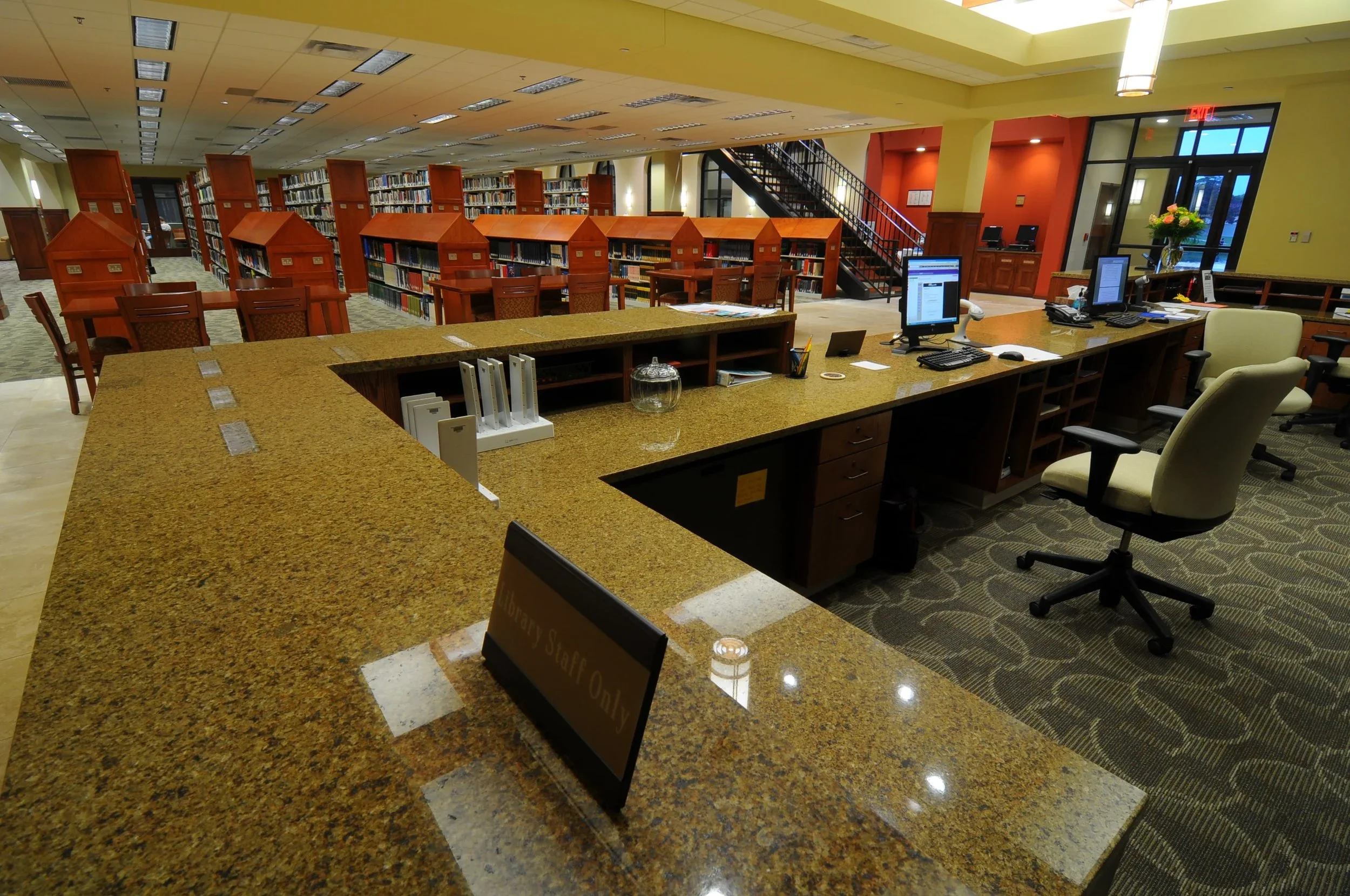 Inside a library showing a front desk with a sign that reads "Library Staff Only," computer workstations, and bookshelves filled with books, with a staircase and sitting area in the background.