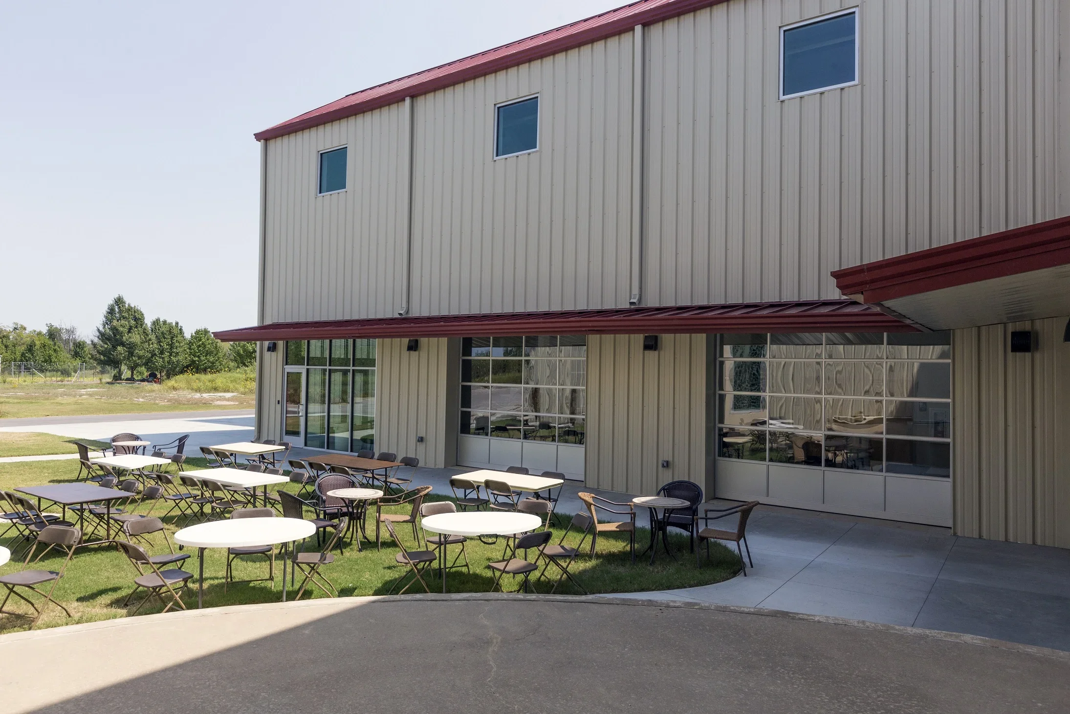 An outdoor patio area with tables and chairs in front of a beige metal building with large windows and a red roof.