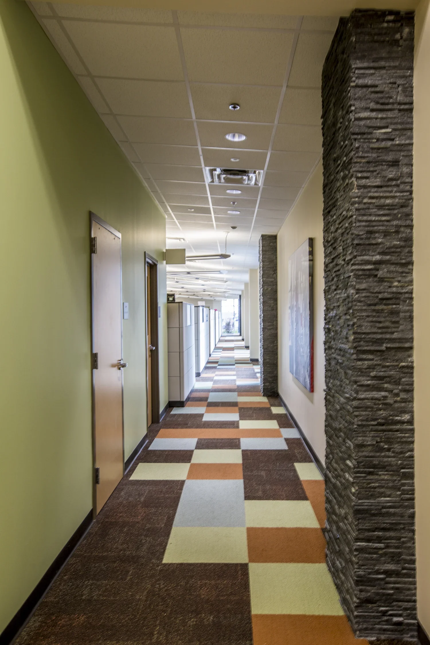 Long brightly lit hallway with multicolored checkered carpet, green and beige walls, and black textured stone columns.