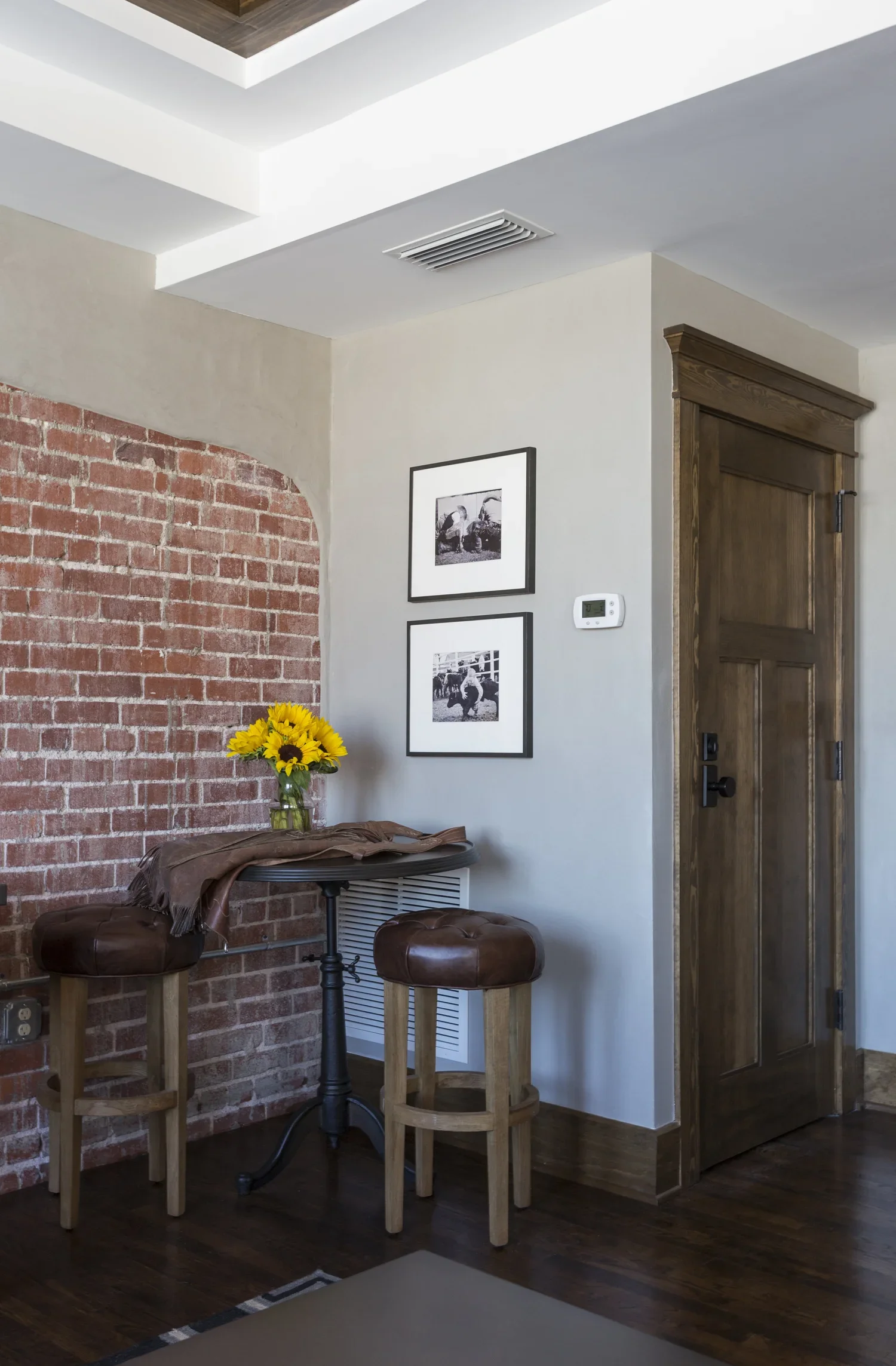 A cozy corner of a room with a brick accent wall, a small black round table with a glass vase of sunflower flowers, and two wooden stools with leather seats. There are black and white framed photographs on the wall near a wooden door, and a thermosta