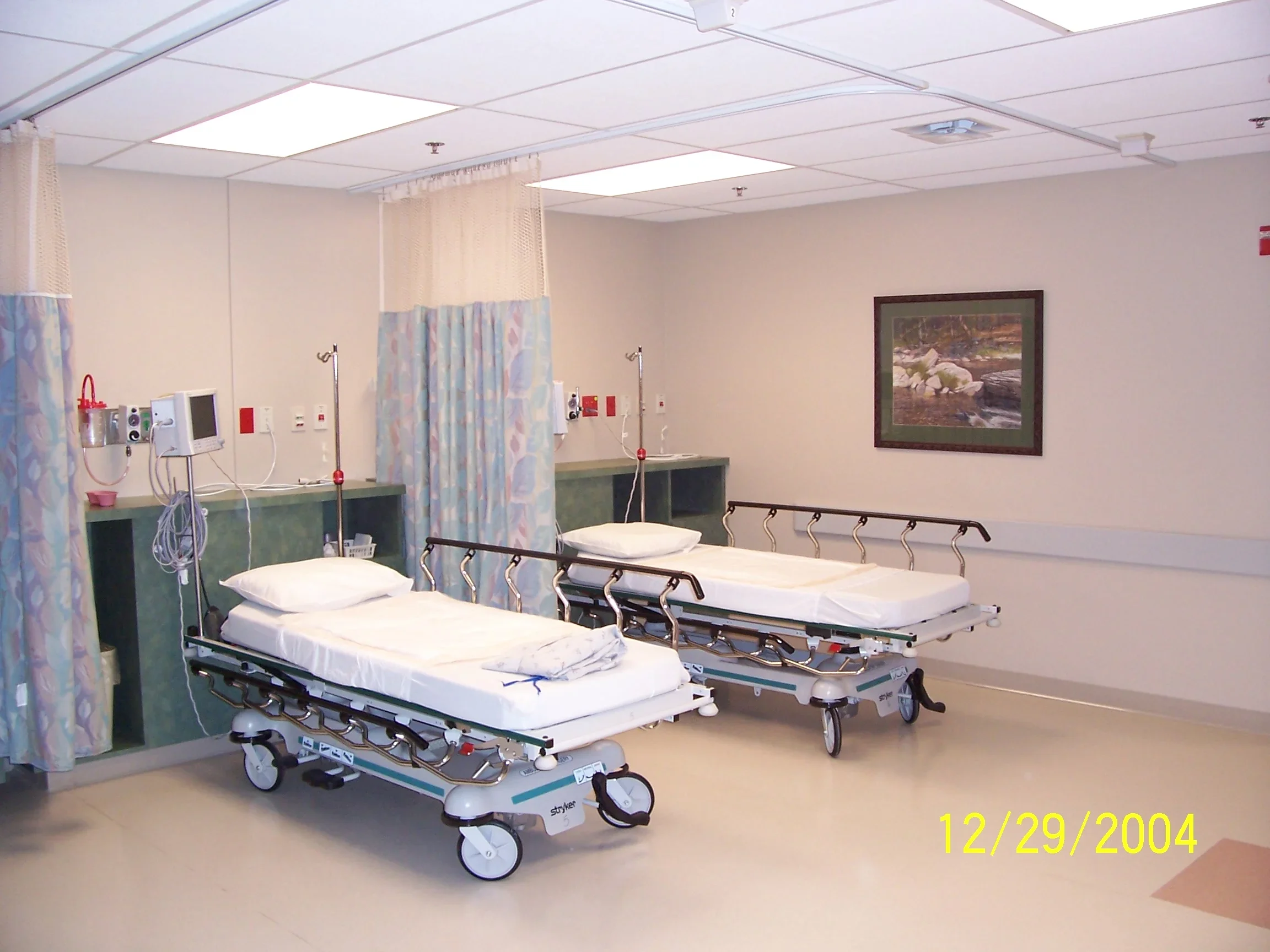 Two empty hospital beds with white sheets and pillows in a clinical room with medical monitors and equipment, curtain dividers, and wall art.