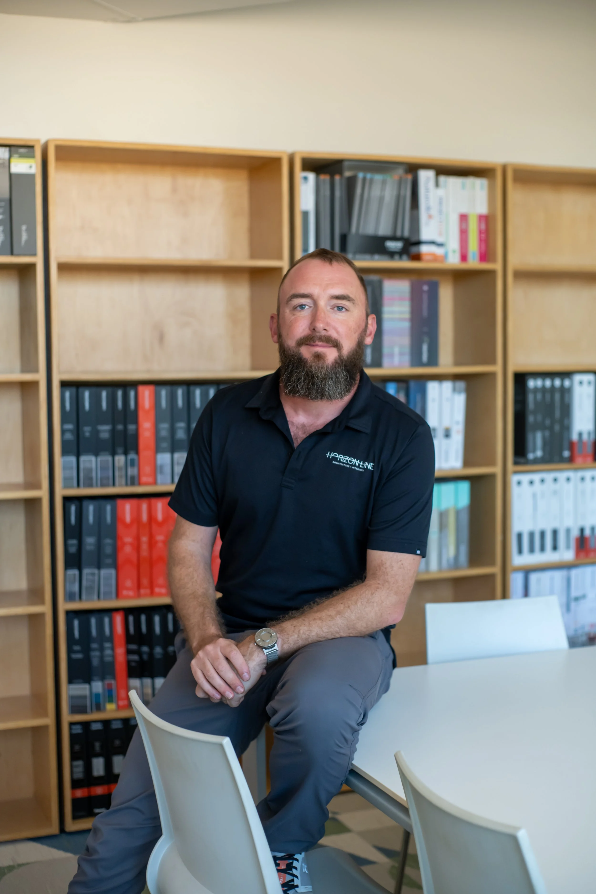 A man with a beard wearing a black polo shirt and gray pants is sitting on the edge of a white table in an office, with bookshelves filled with binders behind him.