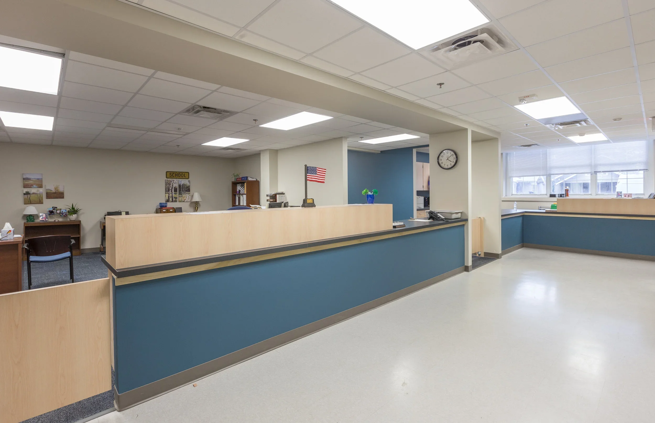 Empty school reception or front desk area with a blue and beige counter, a clock on the wall, American flag, large windows, and various office supplies.