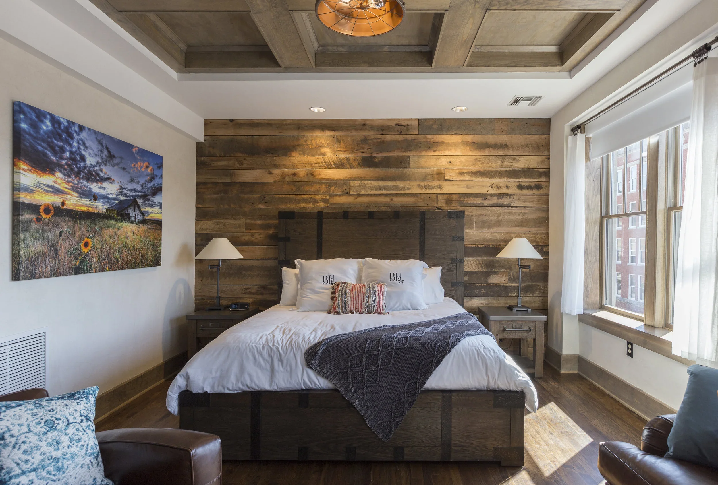 A cozy bedroom with a wooden accent wall behind a bed, white bedding, and two nightstands with lamps. Large windows with white curtains let in natural light, and a landscape painting hangs on the adjacent wall.