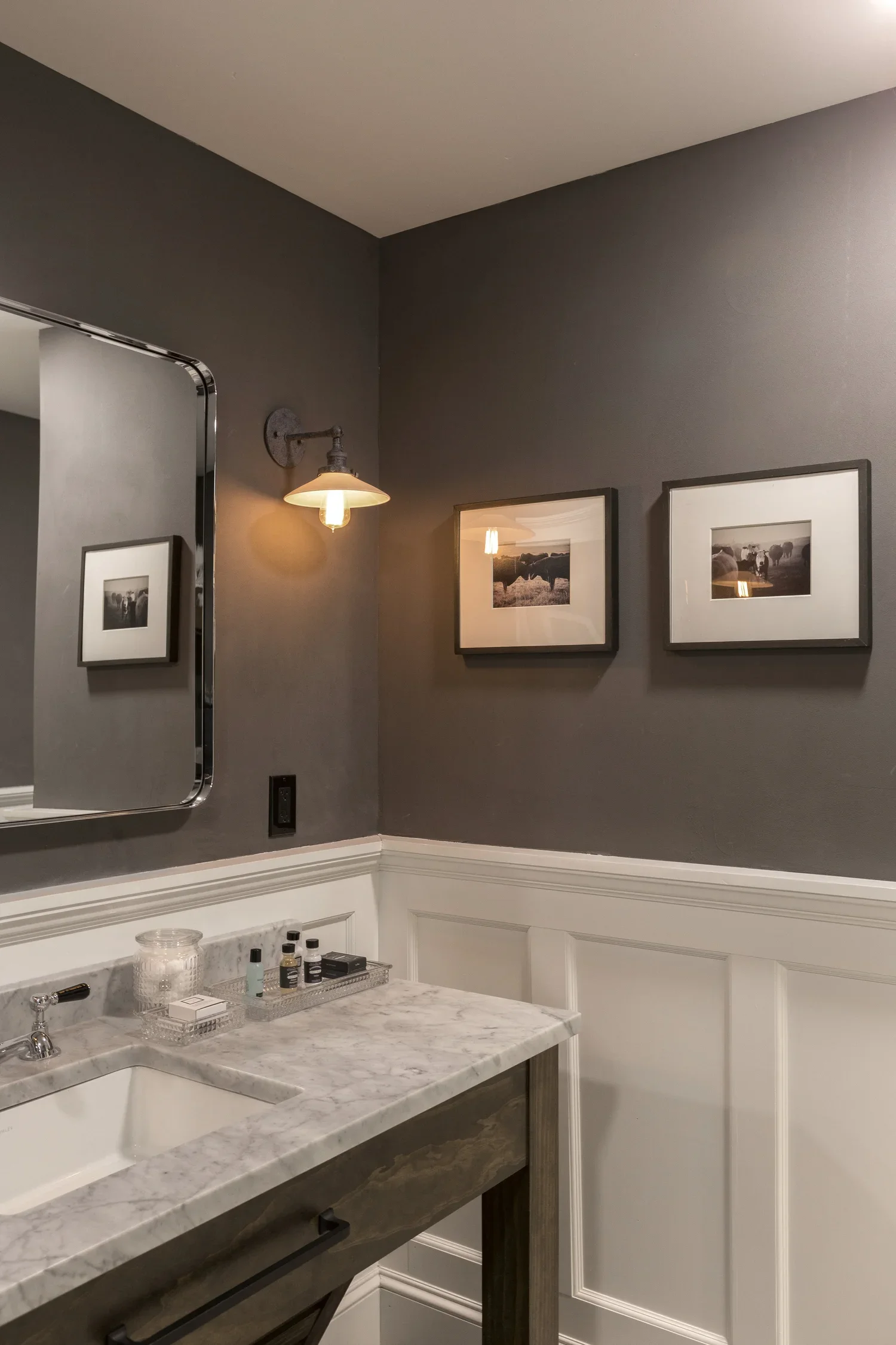 A bathroom with dark gray walls, a marble countertop with toiletries, a rectangular mirror, a wall-mounted light fixture, and two framed photographs of cows and farm scenes.