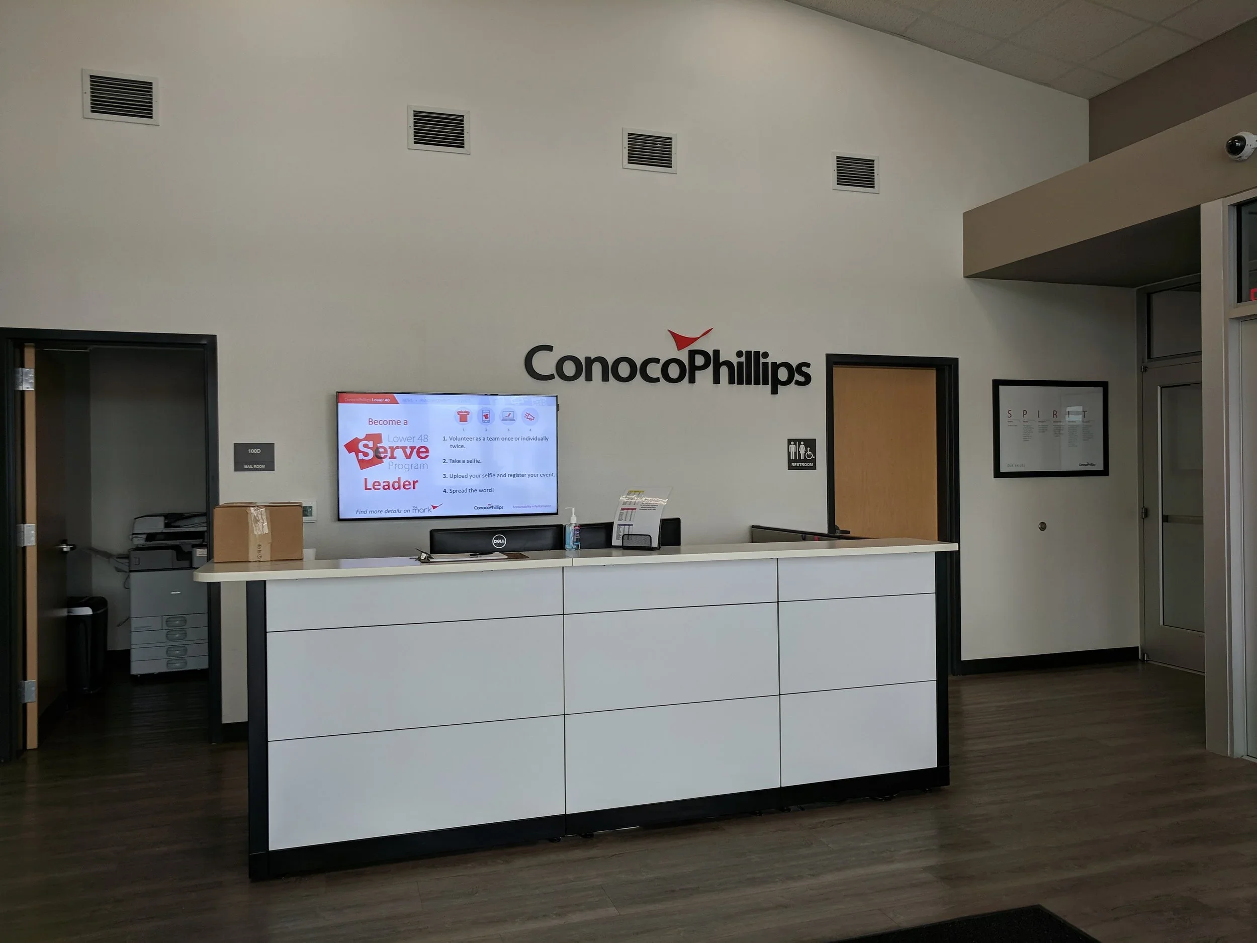 ConocoPhillips lobby reception desk with a wall-mounted screen displaying a volunteer sign-up promotion, and behind the desk is a wall with the ConocoPhillips logo. To the right of the logo is a restroom sign, and there are ceiling vents visible.