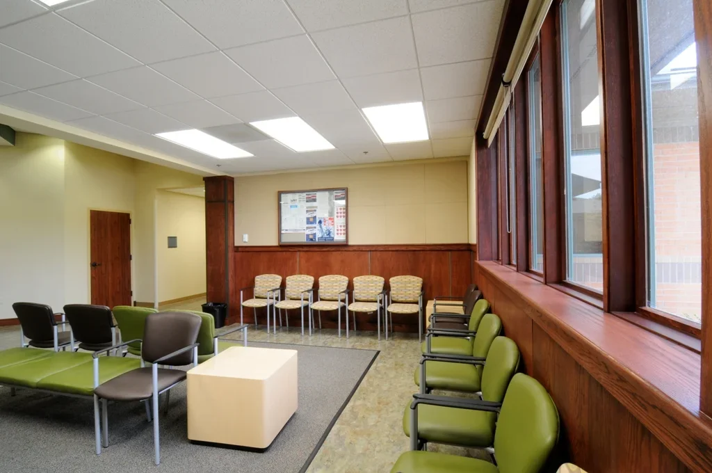 An empty waiting room with green and black chairs, a small table, and a bulletin board on the wall. Large windows on the right side allow natural light.