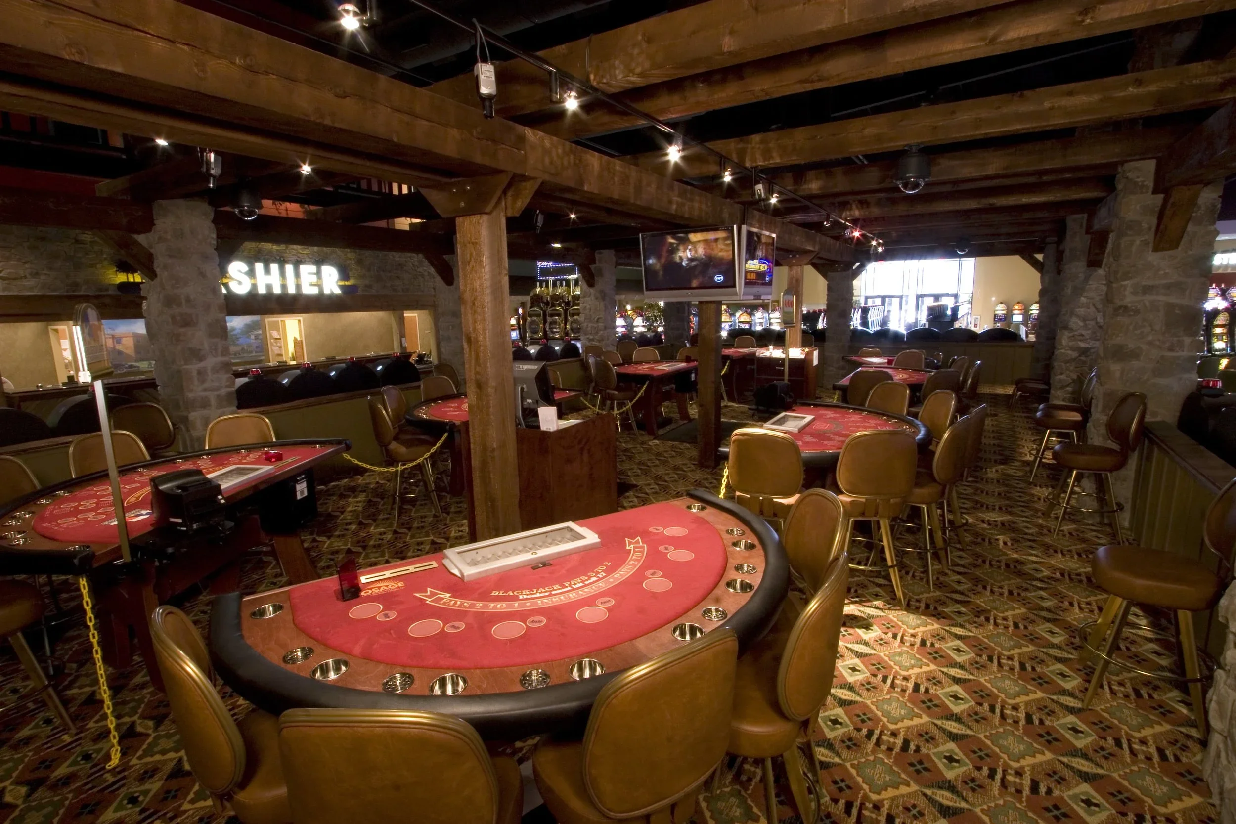 Casino interior with poker tables, slot machines, television screens, stone and wood decor, and ambient lighting.