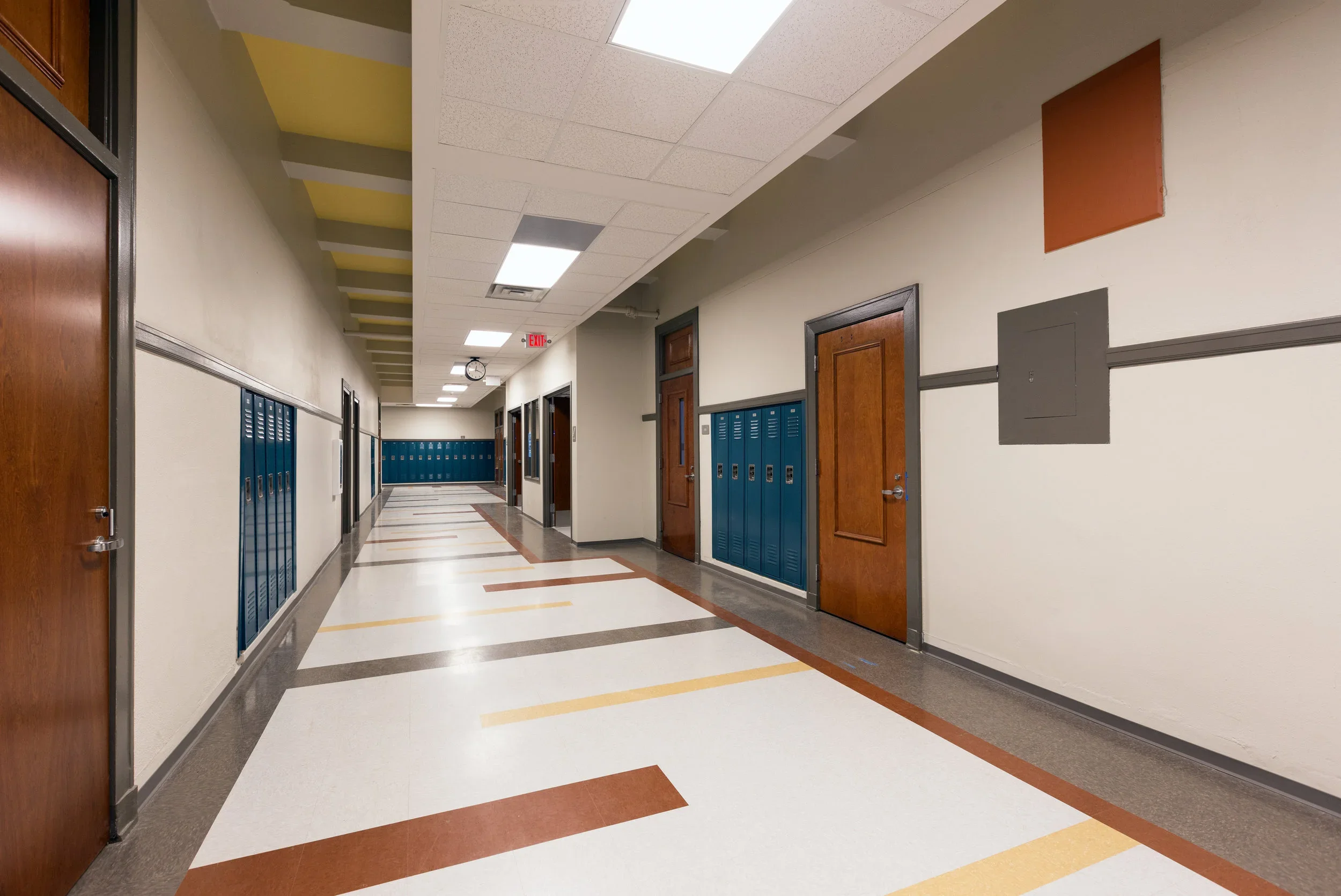 Empty school hallway with blue lockers, brown and beige doors, beige walls, and geometric patterned tile flooring. Fluorescent ceiling lights and a clock visible in the background.