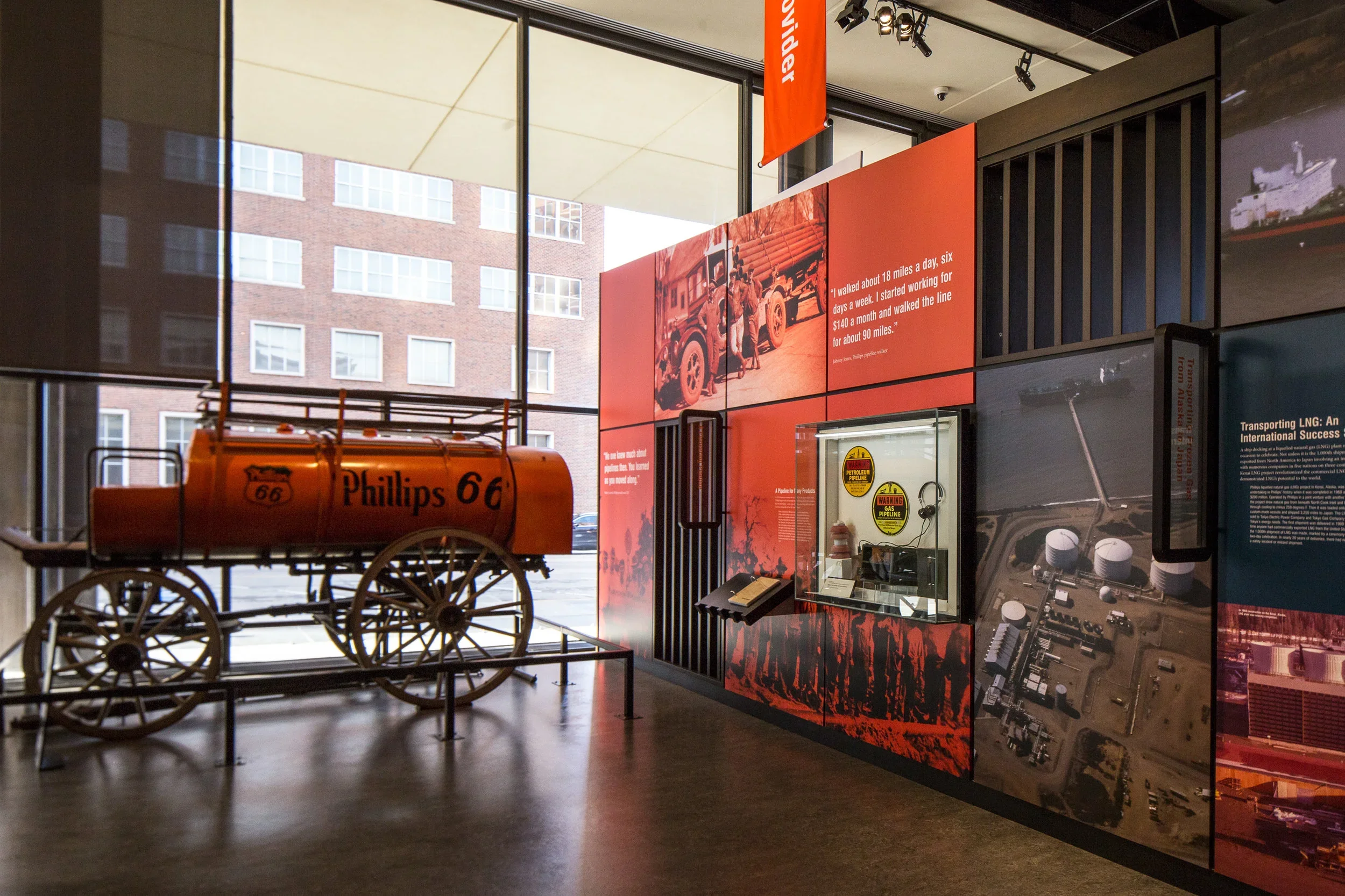 Museum exhibit with a vintage orange Phillips 66 gas car model, informational panels, and a display case, set inside a building with large windows showing a brick building outside.