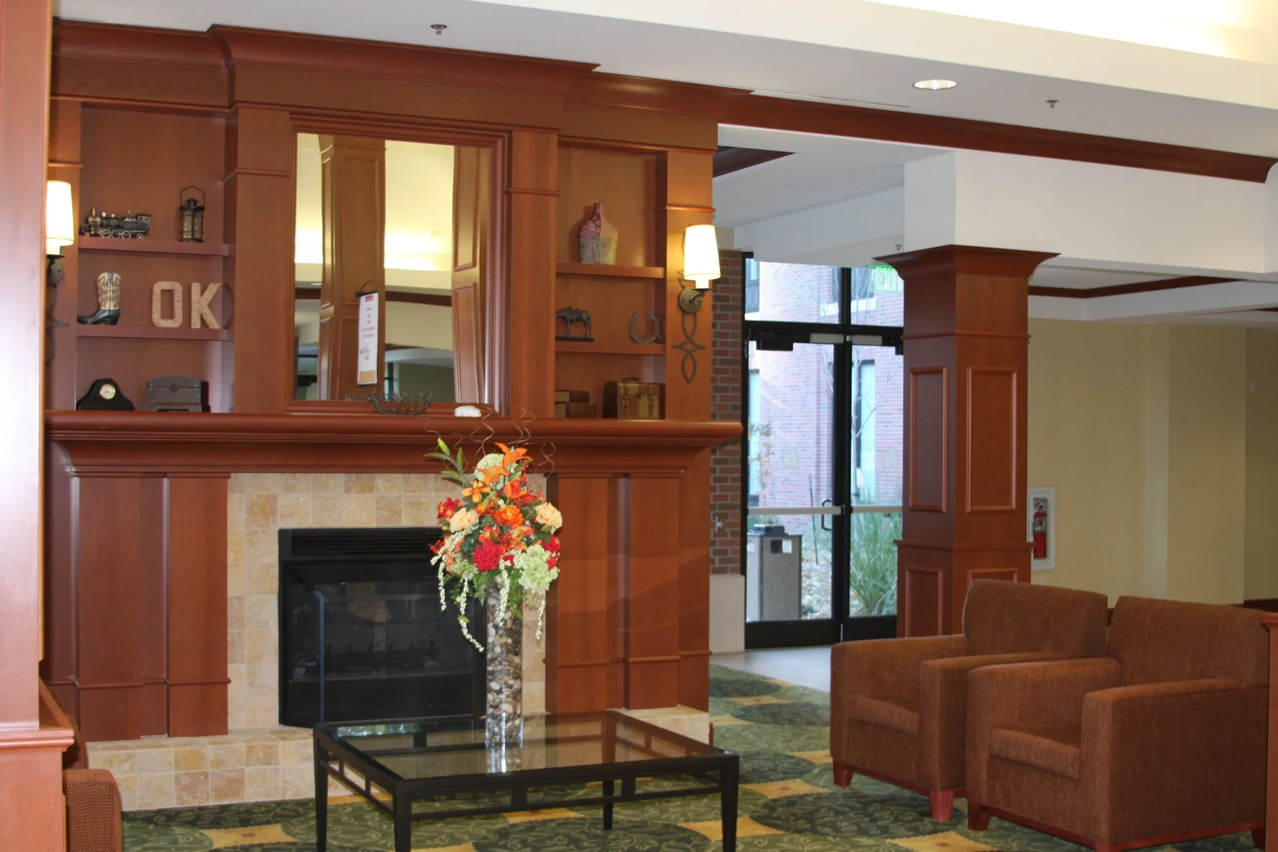 Hotel lobby with wood-paneled wall, fireplace with floral arrangement, sitting area with two brown armchairs, glass-top table, entrance door with glass panels, and decorative items on the shelves.
