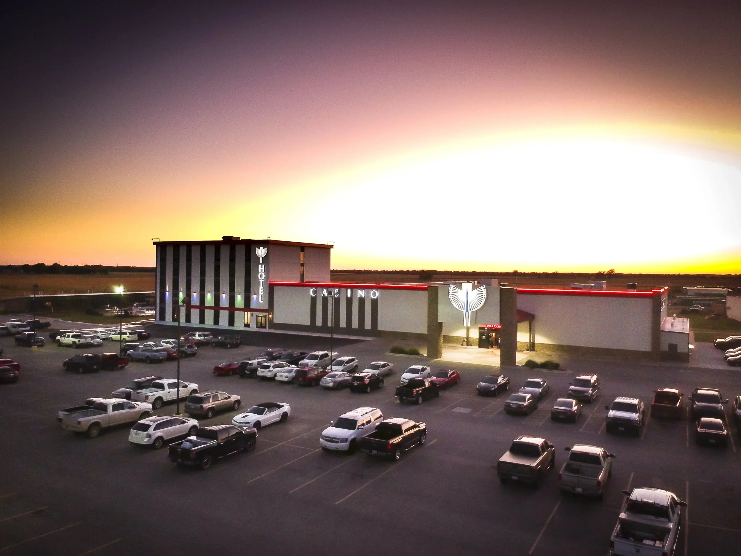 A large parking lot in front of Tonkawa Casino at sunset. The building has a vertical sign that reads 'HOTEL' and 'CASINO' with neon lights and a prominent eagle logo. The sky is vibrant with shades of orange, pink, and purple.
