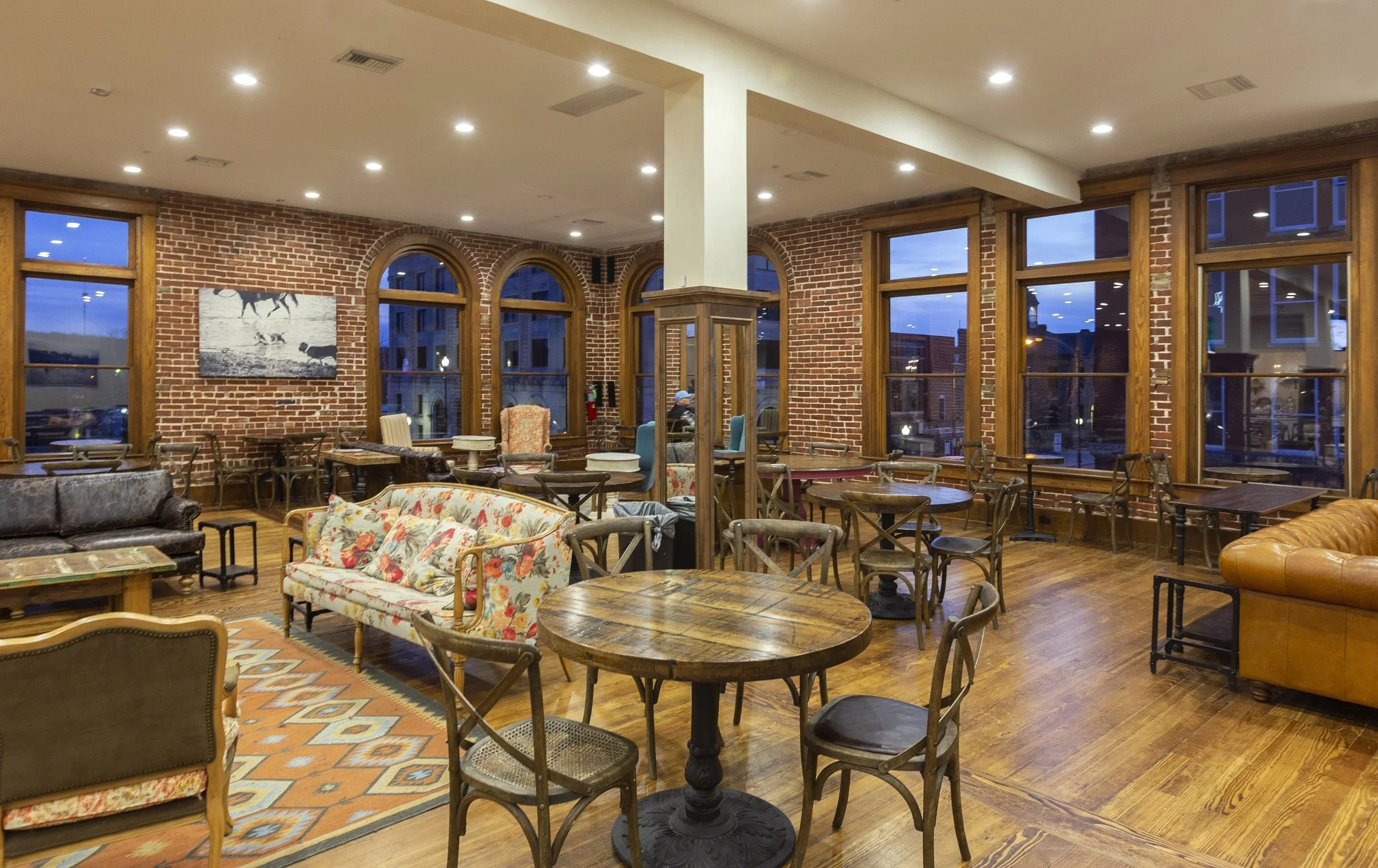 Interior of a cozy cafe with brick walls, large wooden-framed windows, and wooden furniture including tables and chairs. There is a floral sofa, a leather armchair, and colorful artwork on the wall. The space is warmly lit with recessed lighting and 