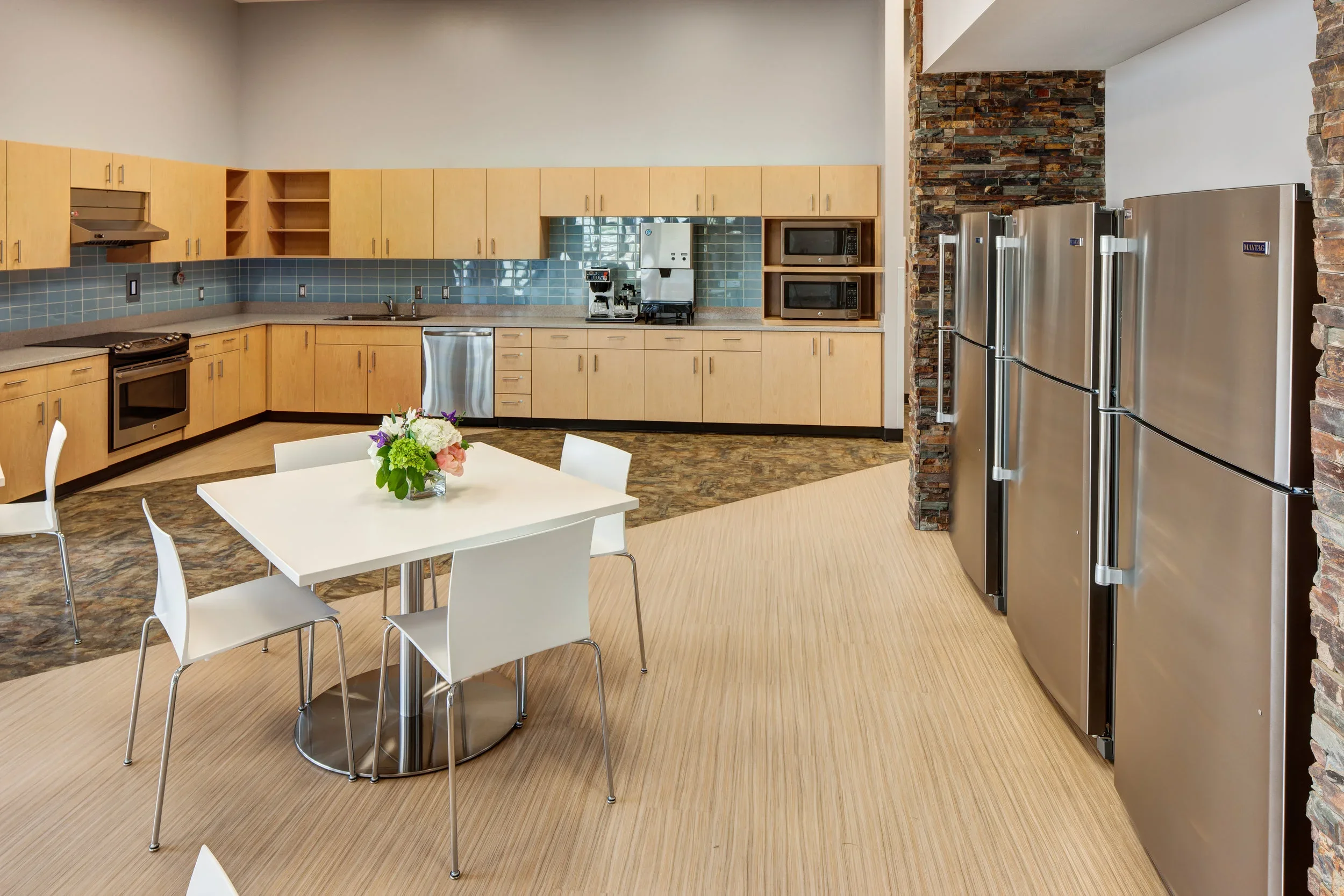 Modern commercial kitchen with stainless steel appliances, wooden cabinets, teal tiled backsplash, white table with chairs, and a floral centerpiece.