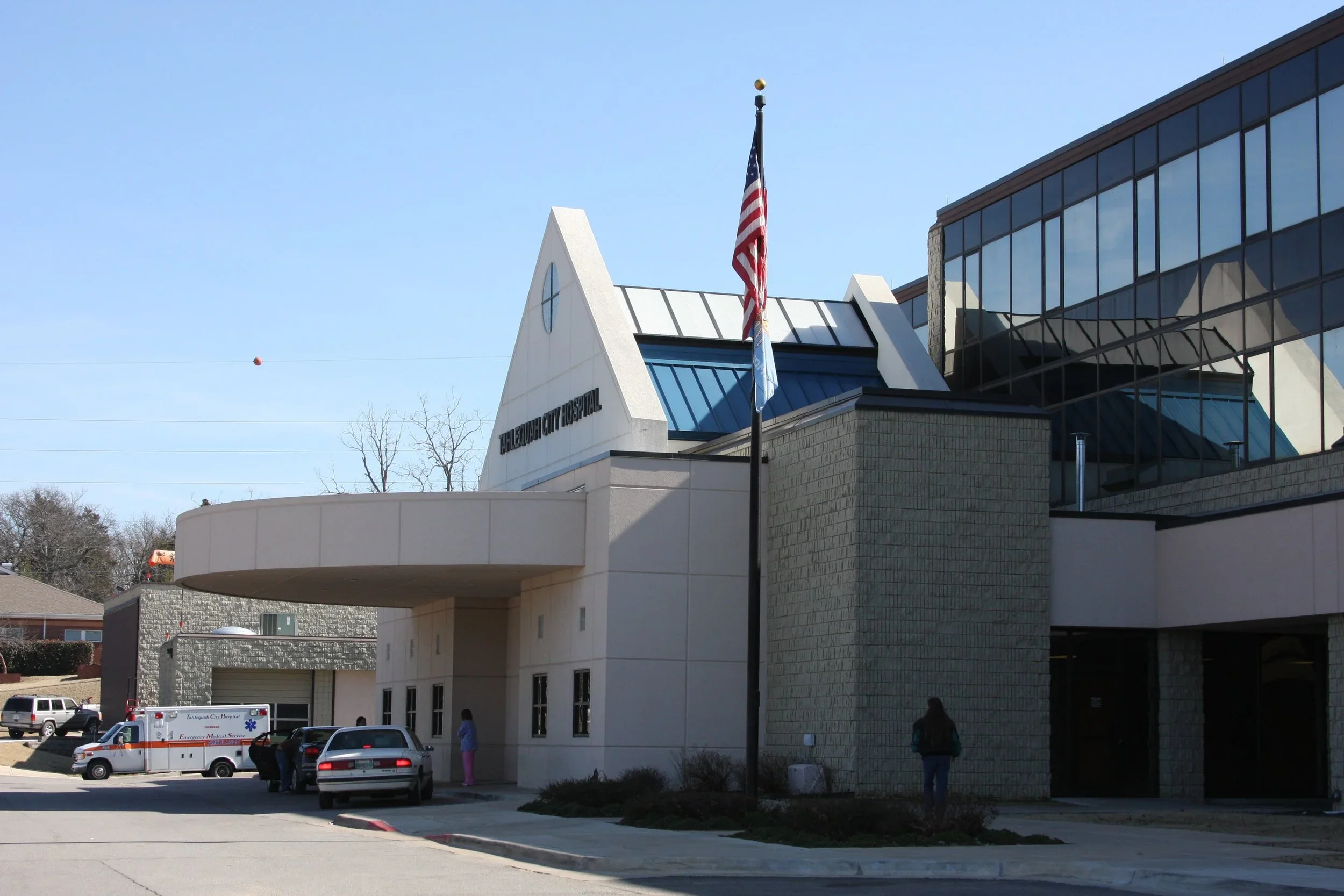 Exterior view of Tahlequah City Hospital with an American flag on a flagpole in front, a few parked cars, and people near the entrance on a clear day.