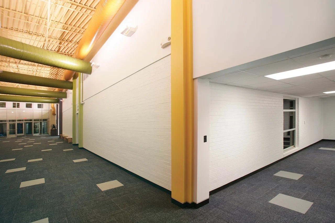 Empty indoor hallway with white brick walls, gray and beige tiled carpet, yellow and green vertical beams, and a ceiling with exposed piping and wooden paneling, illuminated by large ceiling lights.