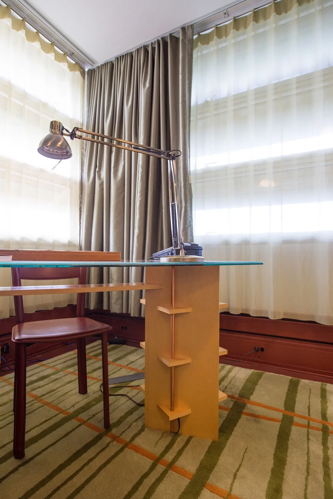 A glass-top desk with a yellow wooden leg and a red wooden chair beside it. There is a silver adjustable desk lamp on the desk. Behind the desk, beige and sheer white curtains cover large windows in a room with a patterned green and orange carpet.