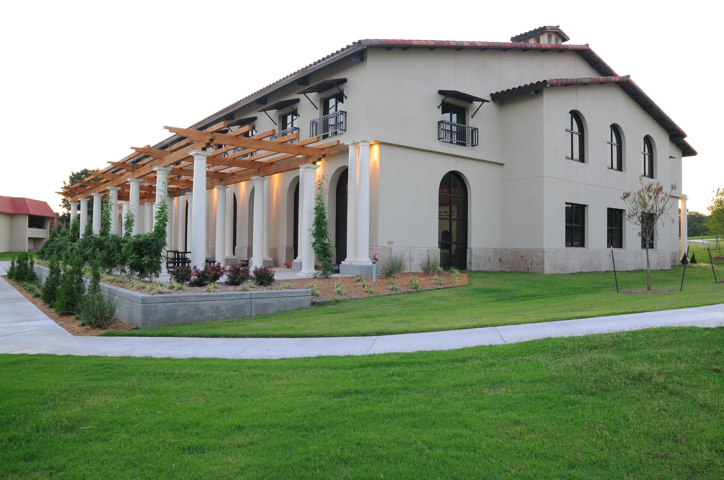 A two-story building with a tiled roof, white exterior walls, arched windows, and a covered porch supported by white columns. The porch has a wooden pergola structure. The building is surrounded by a green lawn and landscaped plants and trees.