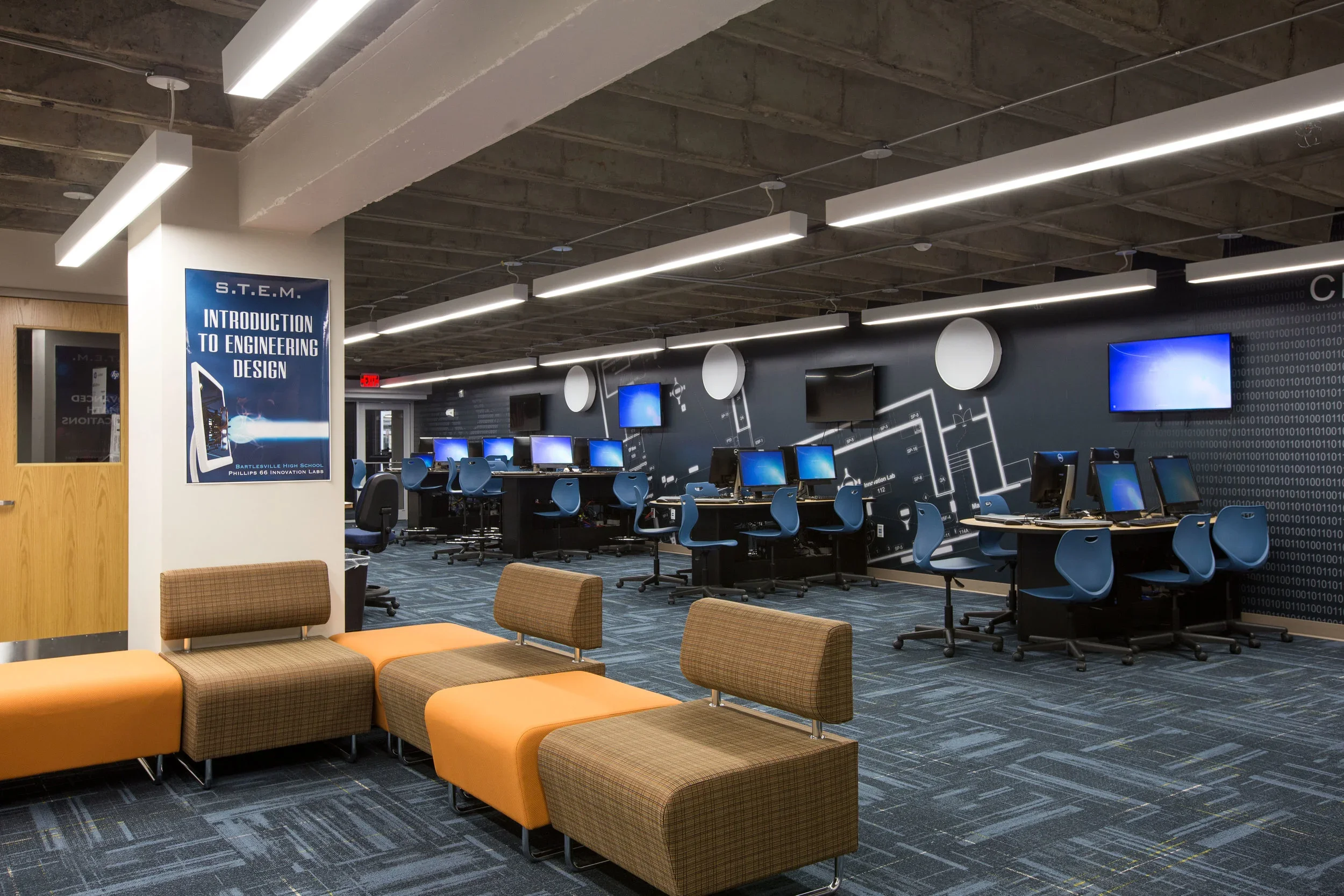 Computer lab with multiple workstations featuring desktop computers and monitors, blue chairs, and a black wall with a white electronic circuit design, illuminated by ceiling lights, with a seating area of orange and brown cushioned benches in the fo