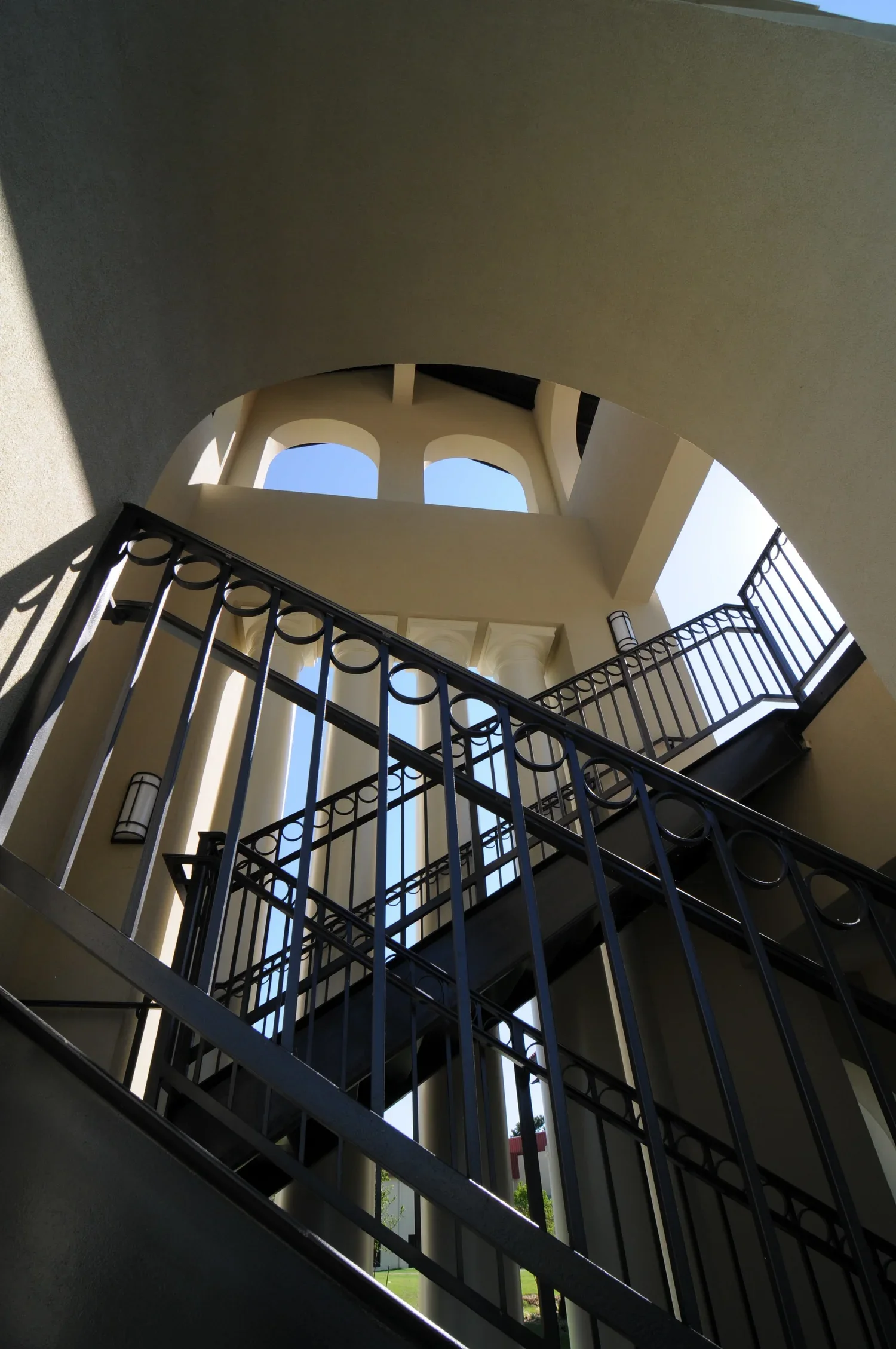 Looking up a staircase and balcony from the ground floor, featuring black metal railings and beige walls, with a view of blue sky through windows.