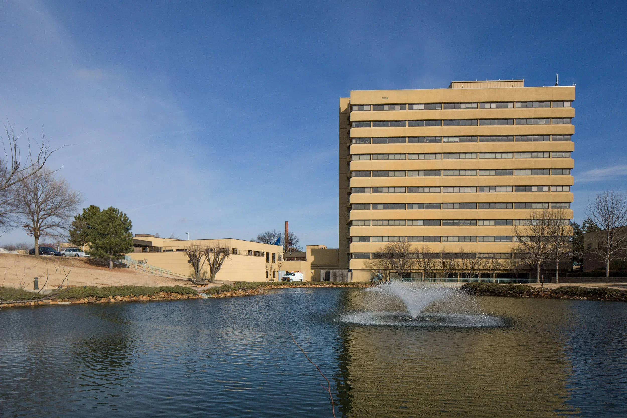 A tall beige office building beside a small body of water with a fountain, surrounded by leafless trees and a few parked cars, under a clear blue sky.