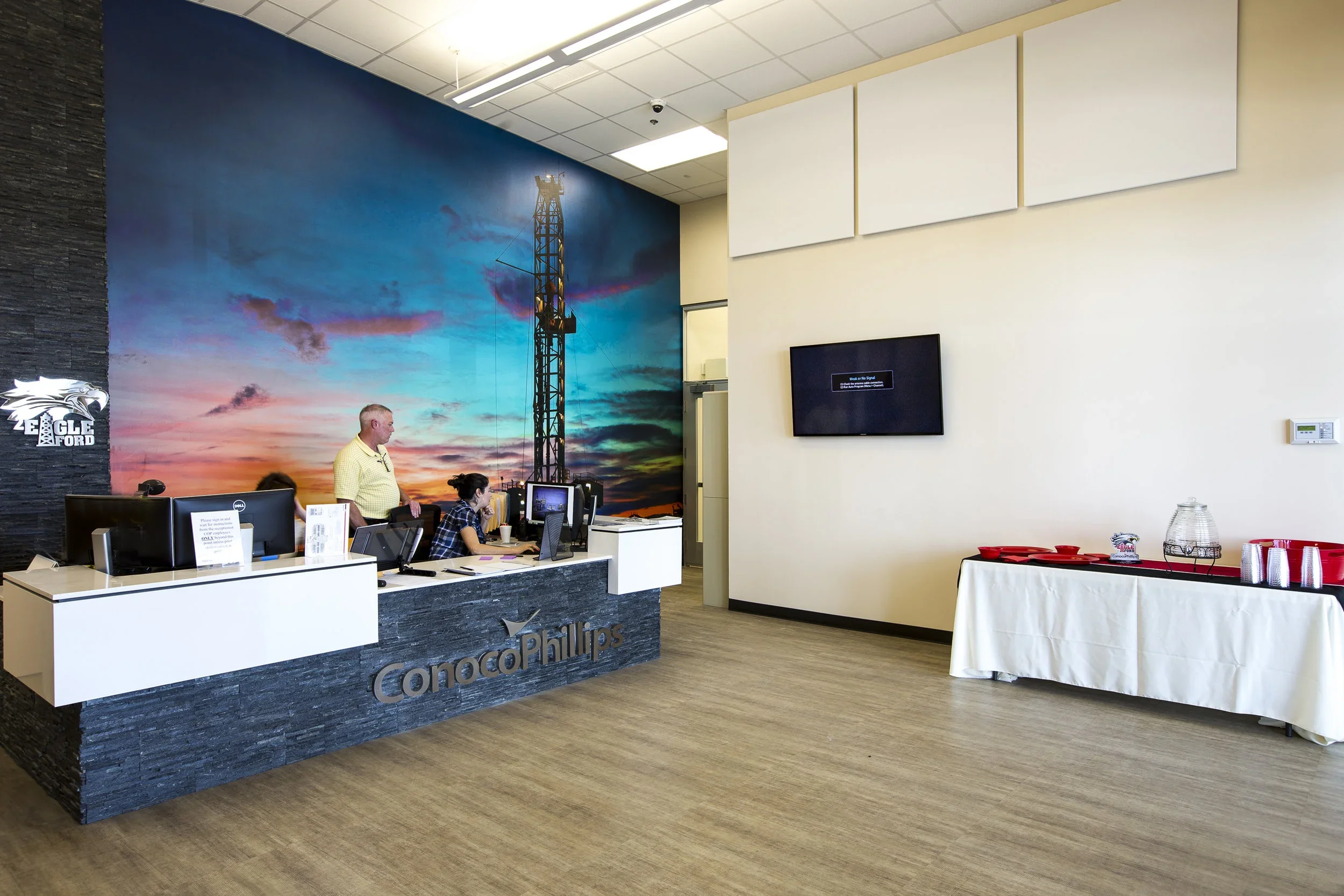 Reception desk at ConocoPhillips with two employees working, and a large mural of an oil drilling rig at sunset on the wall behind. There is a display screen on the wall to the right and a table with refreshments on the far right.