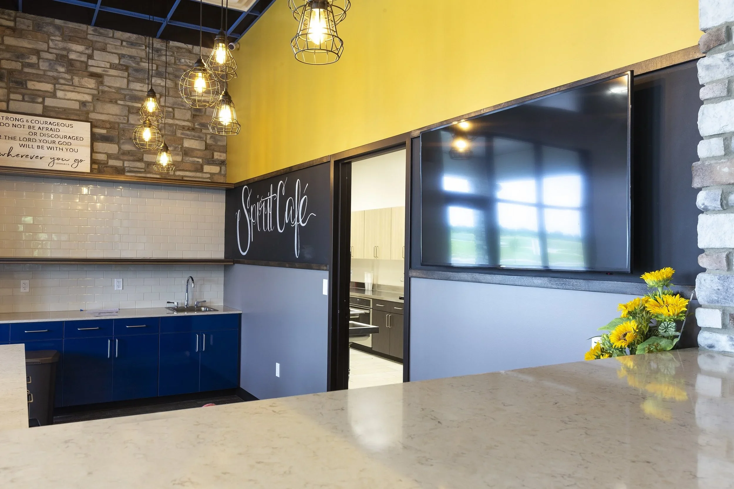 Interior of Spirit Church in Bartlesville, OK with a coffee shop with hanging light fixtures, a yellow accent wall, a blue lower cabinet, a sign that reads 'Spirit Cafe,' a large TV screen, and a bouquet of yellow flowers on a beige countertop.