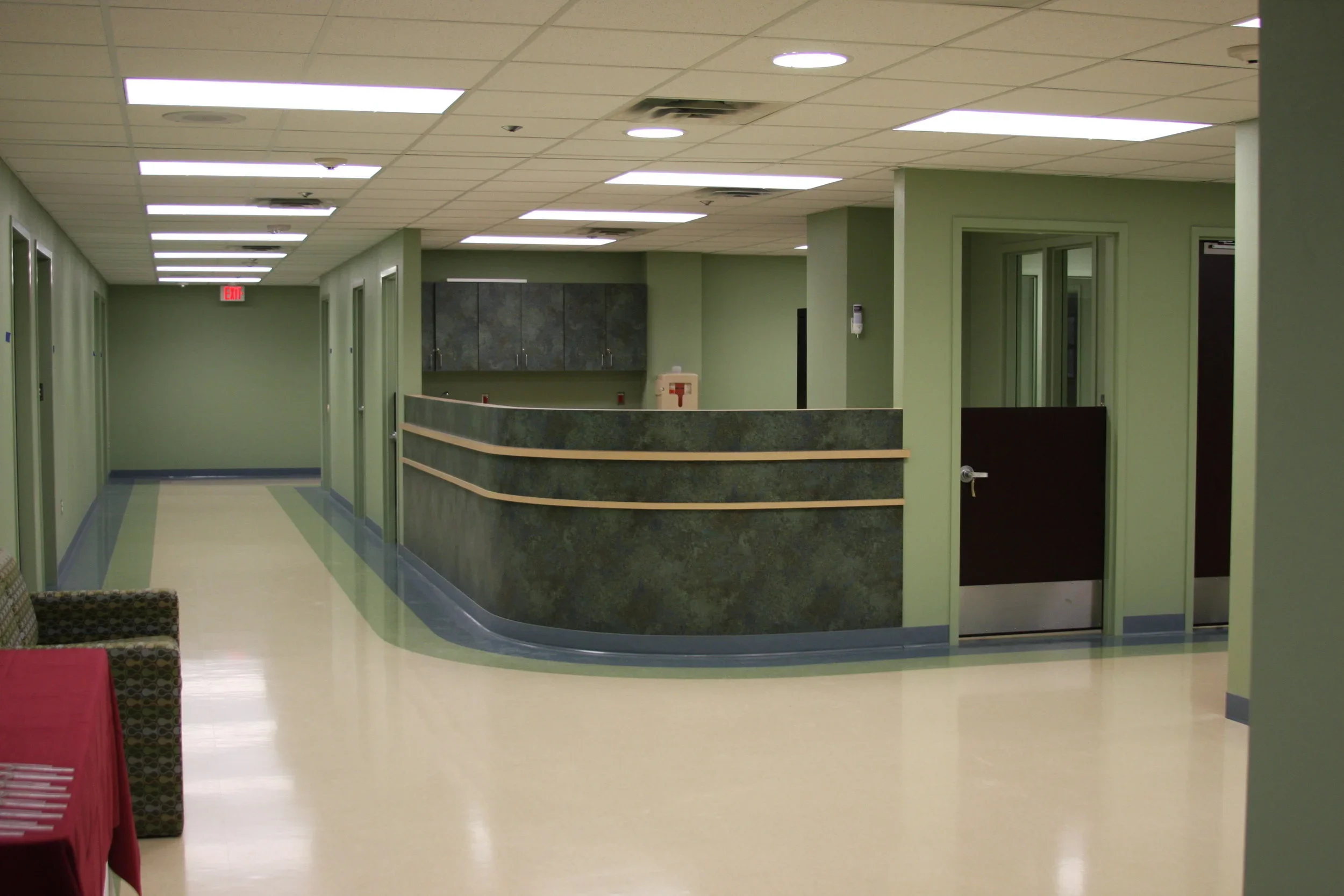Empty hospital reception area with green walls, a curved reception desk, and a hallway with closed doors.