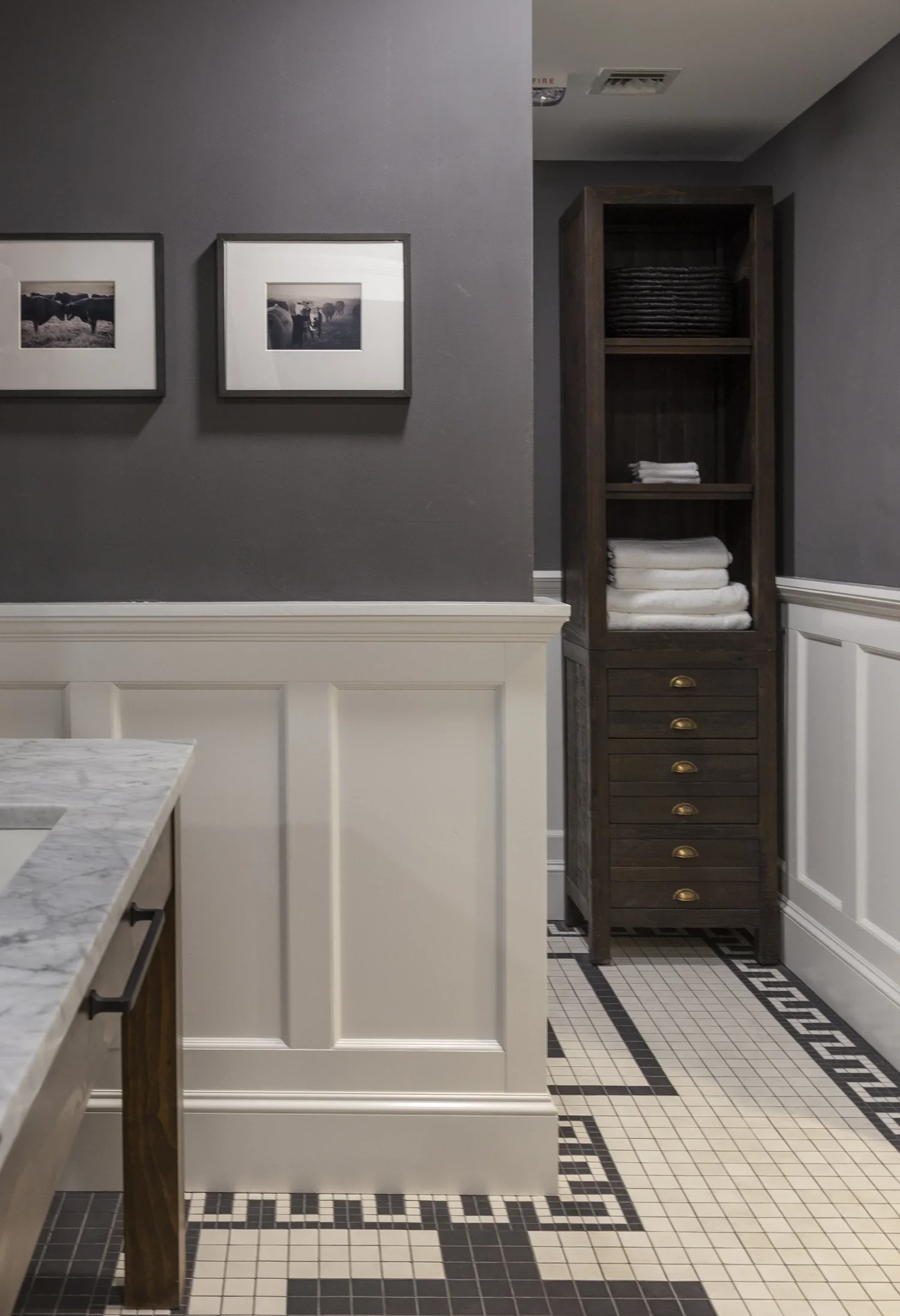 A corner of a room with dark gray and white walls, two framed black-and-white photographs, a wooden cabinet with shelves holding folded towels, and a black and white tiled floor.