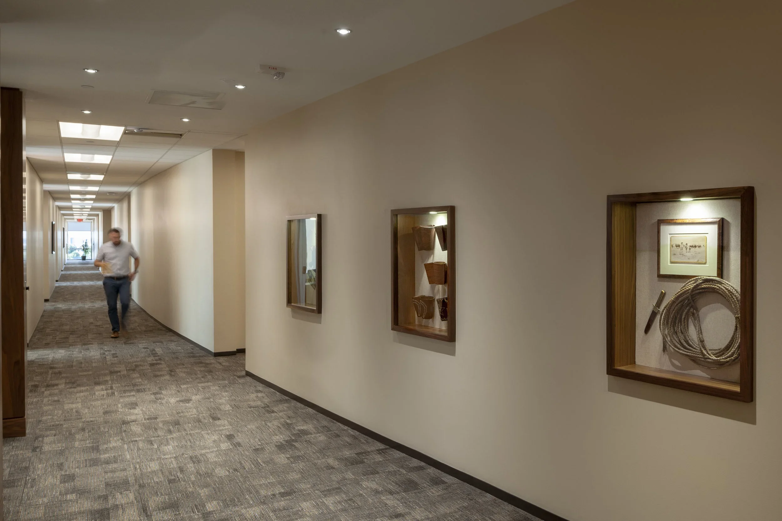 A man walking down a long, well-lit hotel hallway with framed artwork and decorative display cases on the walls.