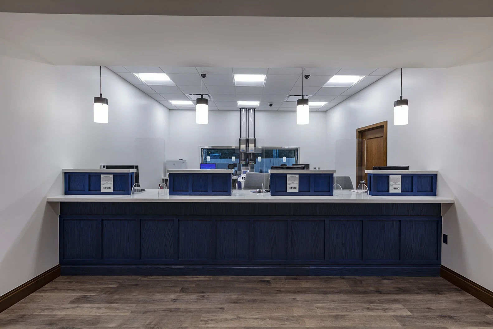 Bank teller area with four desks, blue dividers, pendant lights, wooden flooring, and a window in the background.