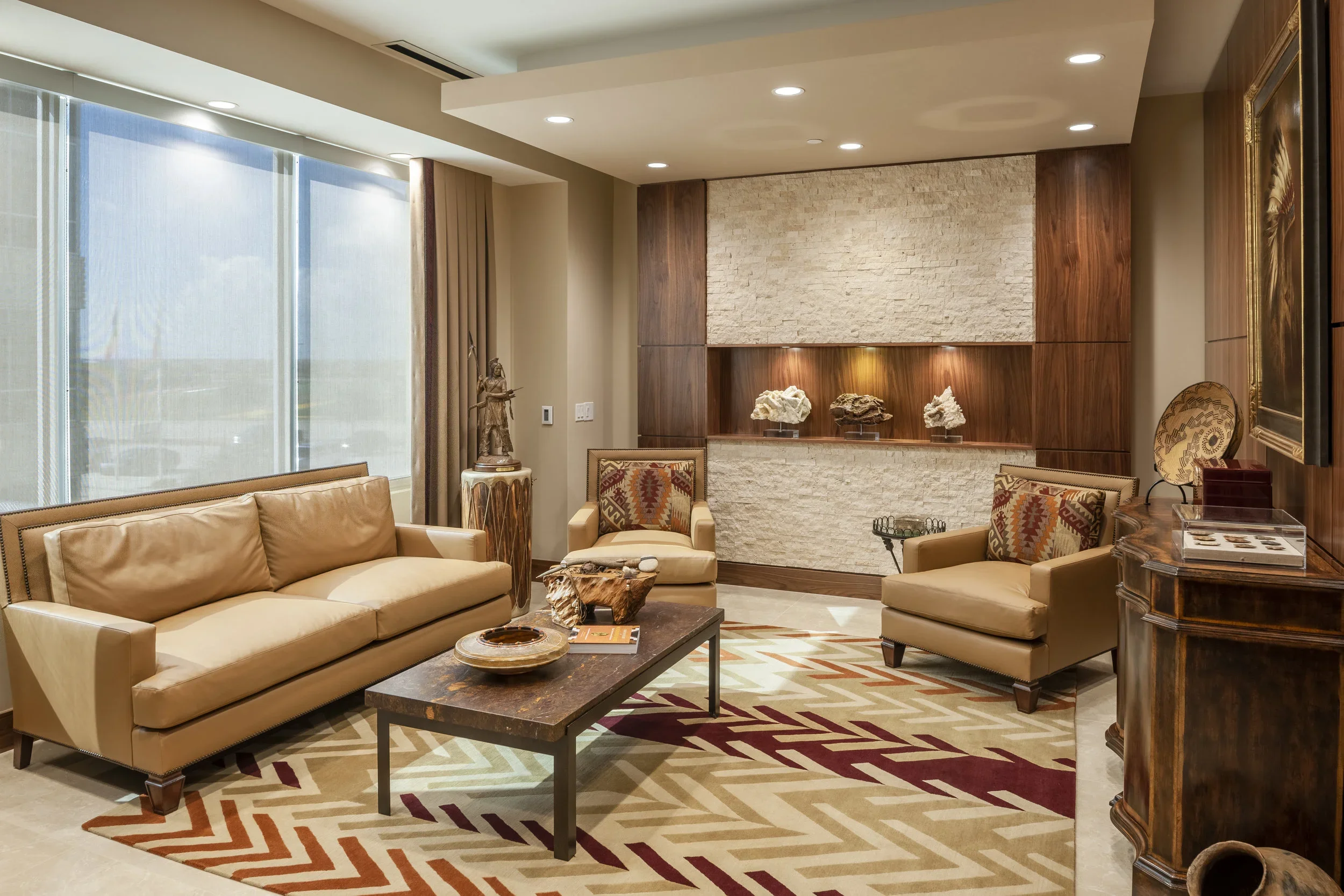 A modern living room with beige leather sofa and armchairs, a wooden coffee table, and decorative objects on a colorful geometric rug. Large window with blinds, wall featuring textured stone and wood panels, with art pieces on display.