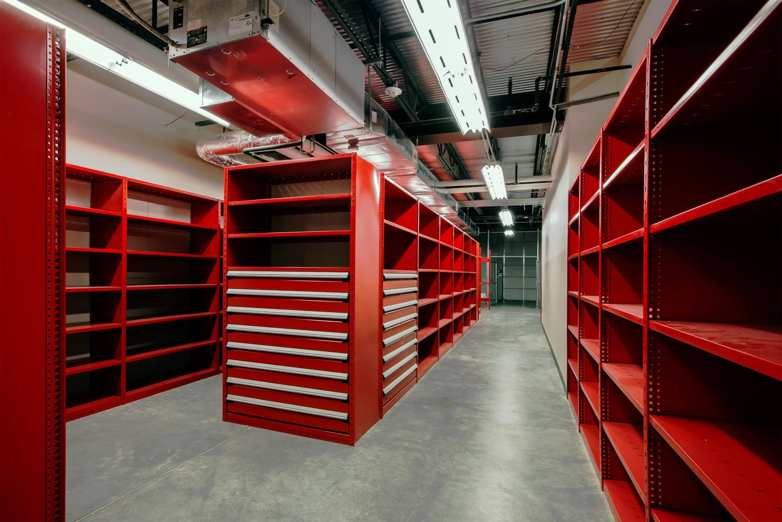 Empty red metal shelving units in a hardware store or warehouse aisle, with overhead fluorescent lighting and exposed ceiling ductwork.