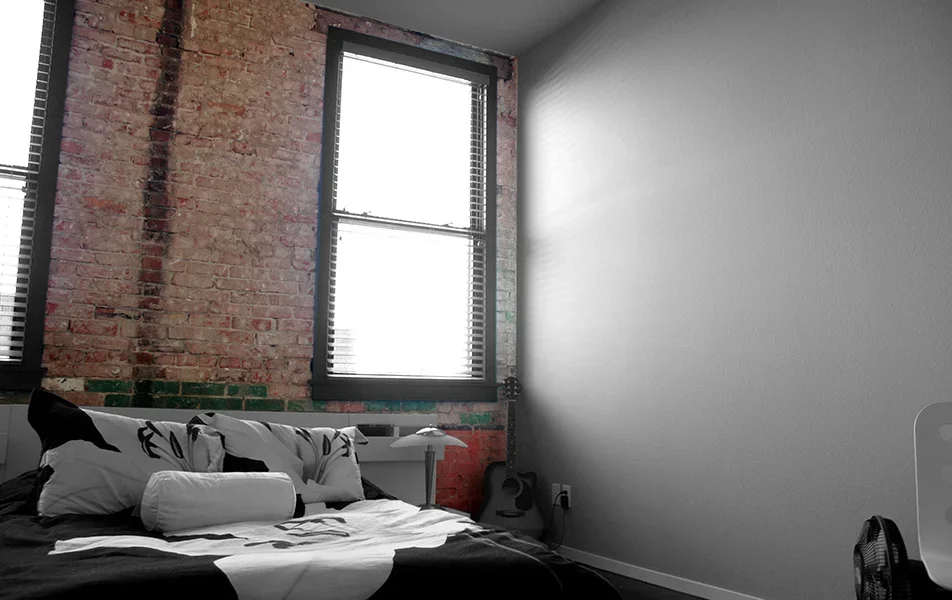 A bedroom with an exposed brick wall, a large window with blinds, a bed with black and white bedding, a white pillow, a white bedside lamp, and an acoustic guitar leaning against the wall.