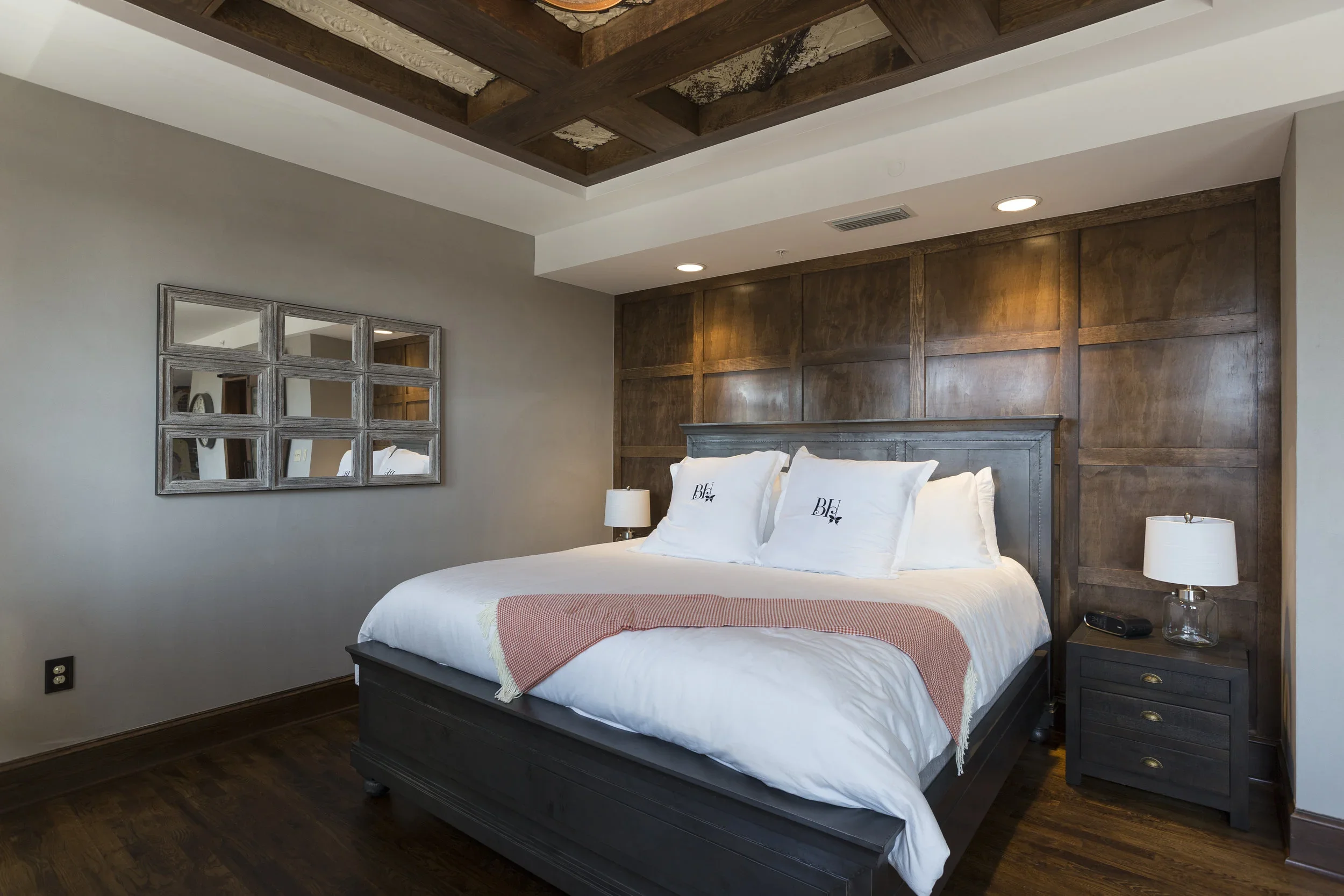 A bedroom with a dark wood bed frame, white bedding, and pillows embroidered with 'BH.' There are matching nightstands with glass lamps and a mirror on the wall. The wall behind the bed has wood paneling, and the ceiling has a decorative wooden desig