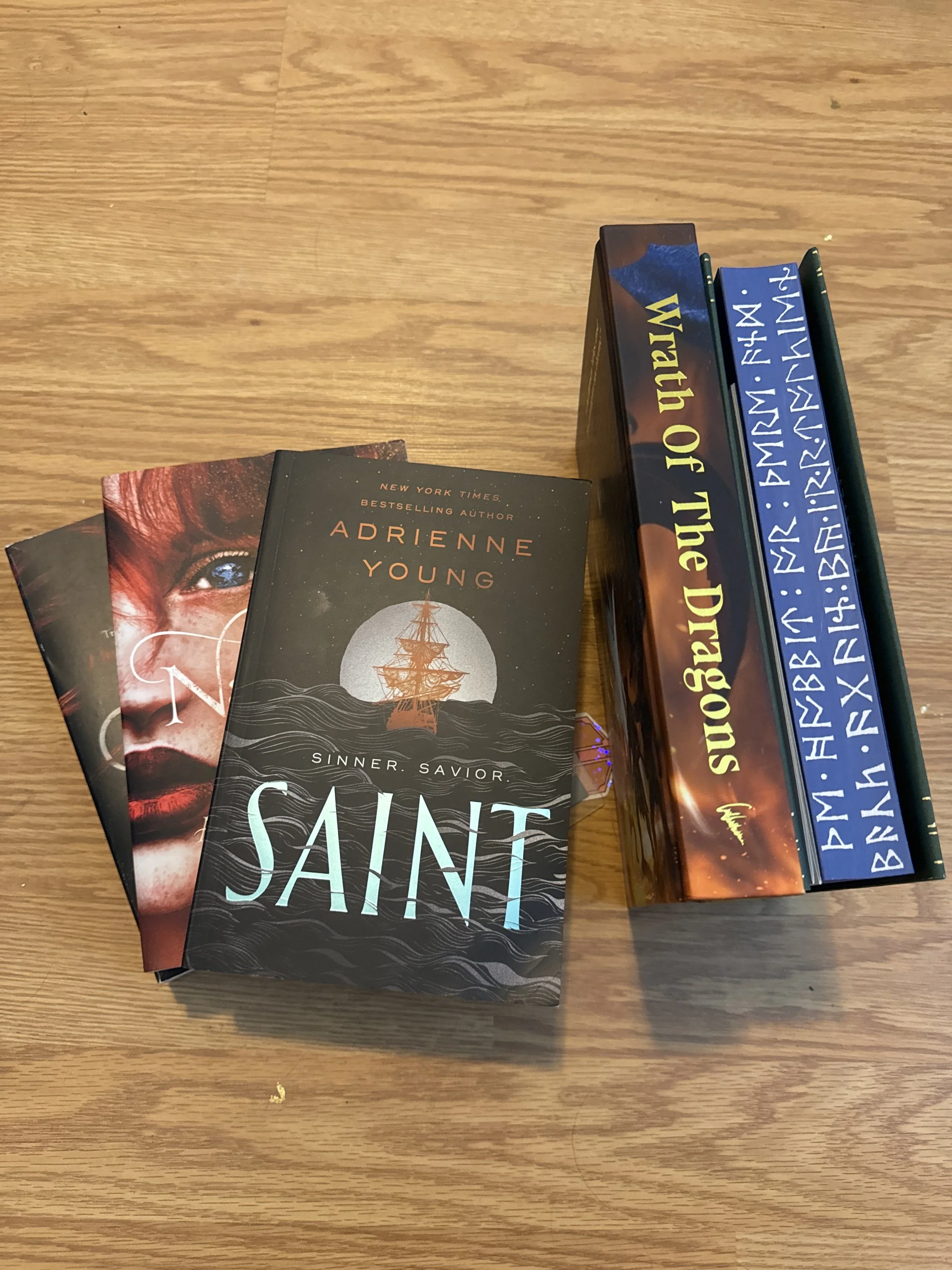 Photograph of several books on a wooden table, including 'Saint' by Adrienne Young, 'The Watch of the Night' and other titles.