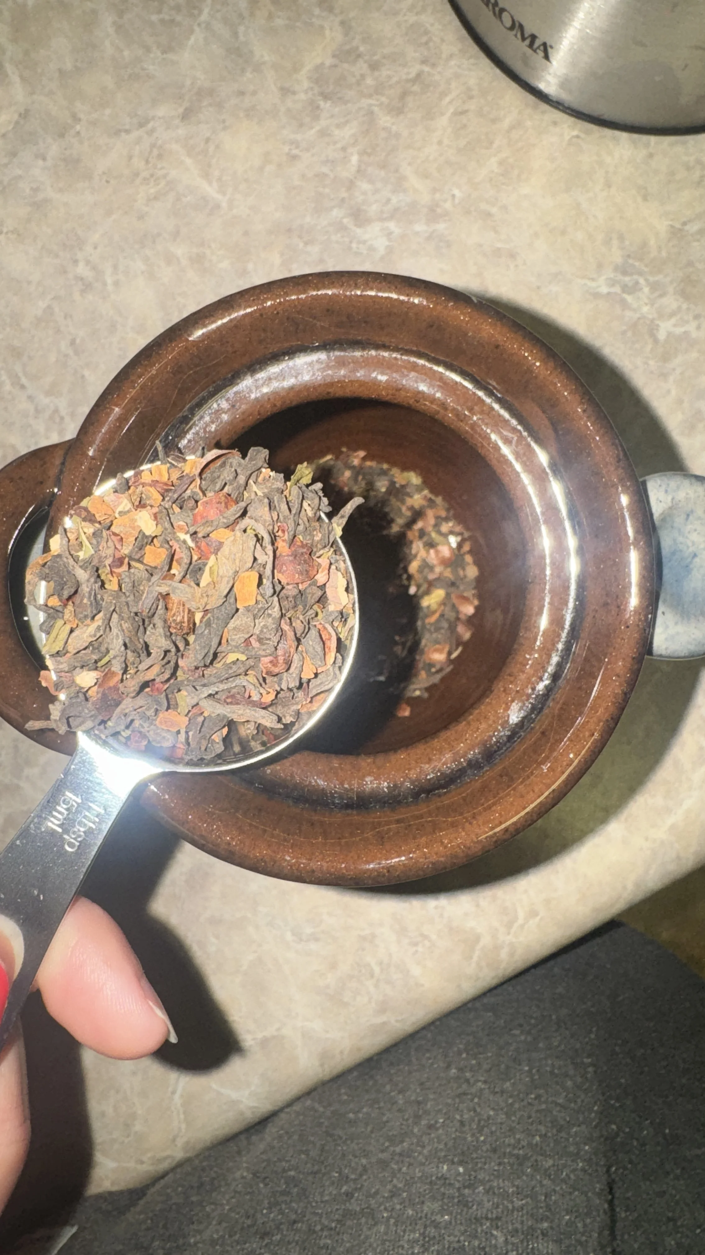 A metal measuring spoon filled with dried loose leaf tea being held over an open ceramic teapot.