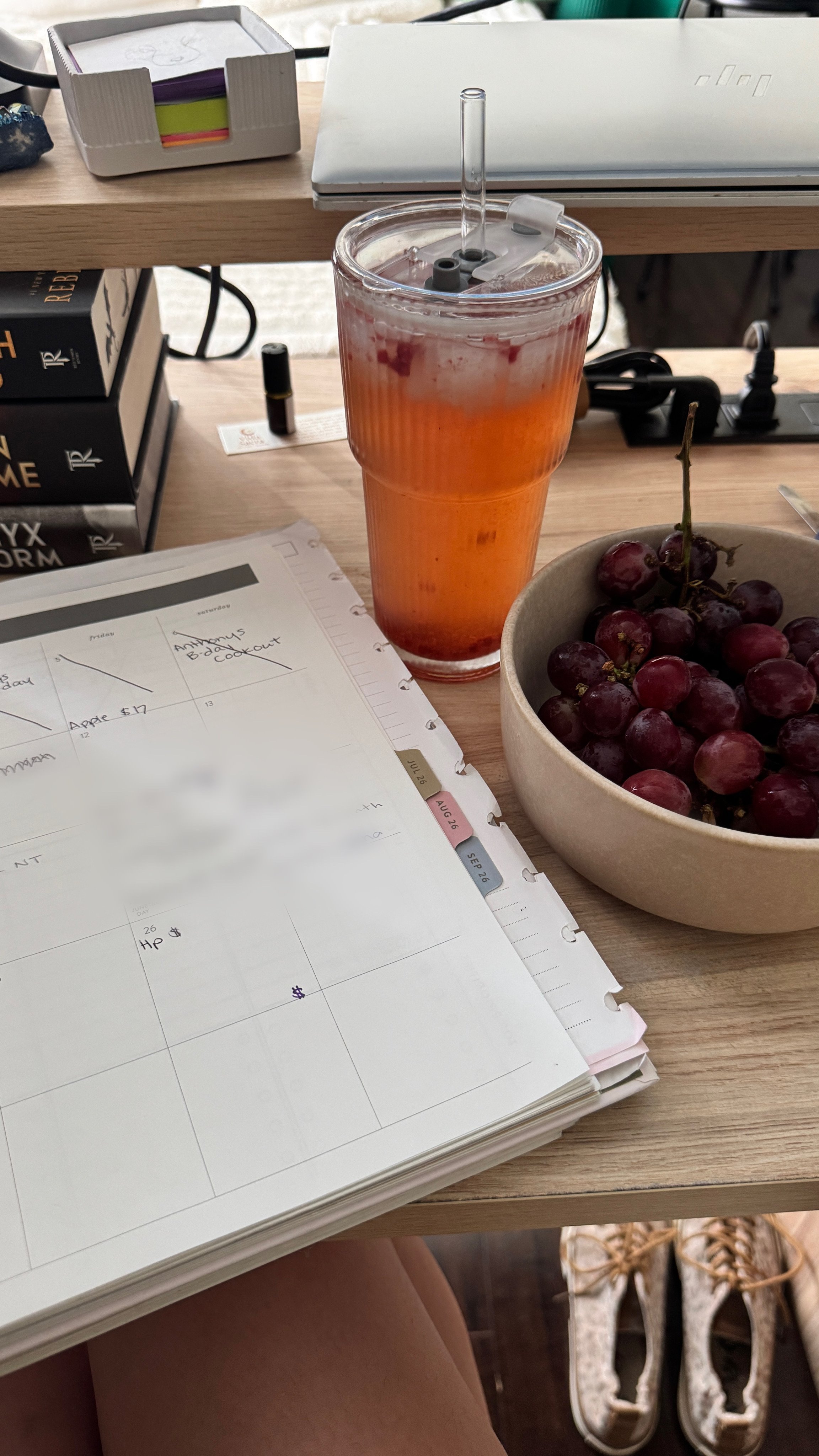 A desk with a stack of books, a planner, a laptop, a notebook, a bowl of grapes, and a tall glass of what appears to be iced tea or a fruit beverage. Shoes are visible below the desk.