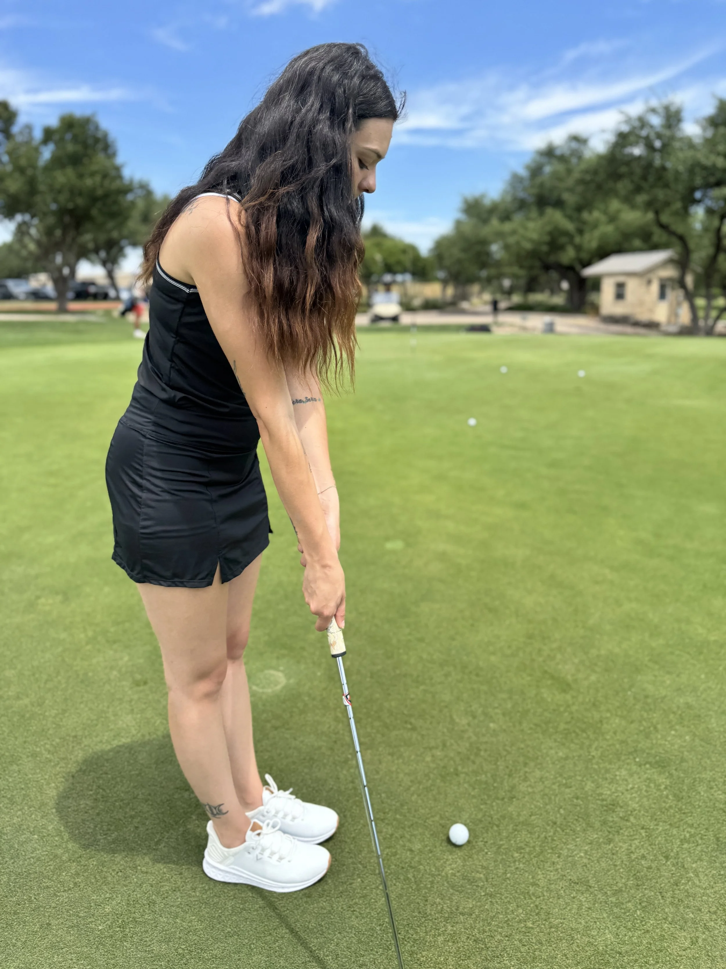 A woman with long dark hair stands on a golf course, preparing to putt a golf ball. She is wearing a black athletic dress and white sneakers, with tattoos on her arm and ankle visible. The background features trees, a building, and a blue sky.