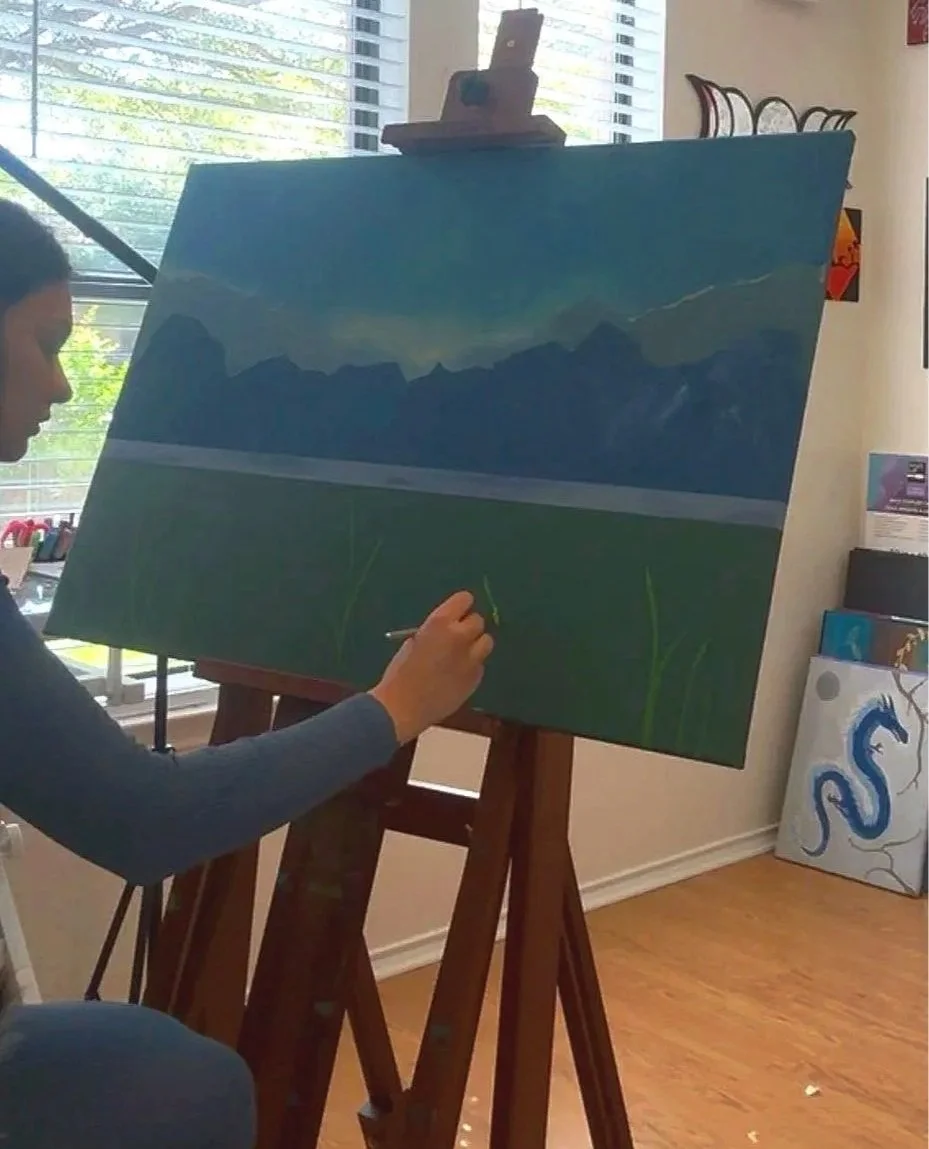 An artist painting a landscape on a large canvas set on an easel, with mountains in the background and a grassy field in the foreground.
