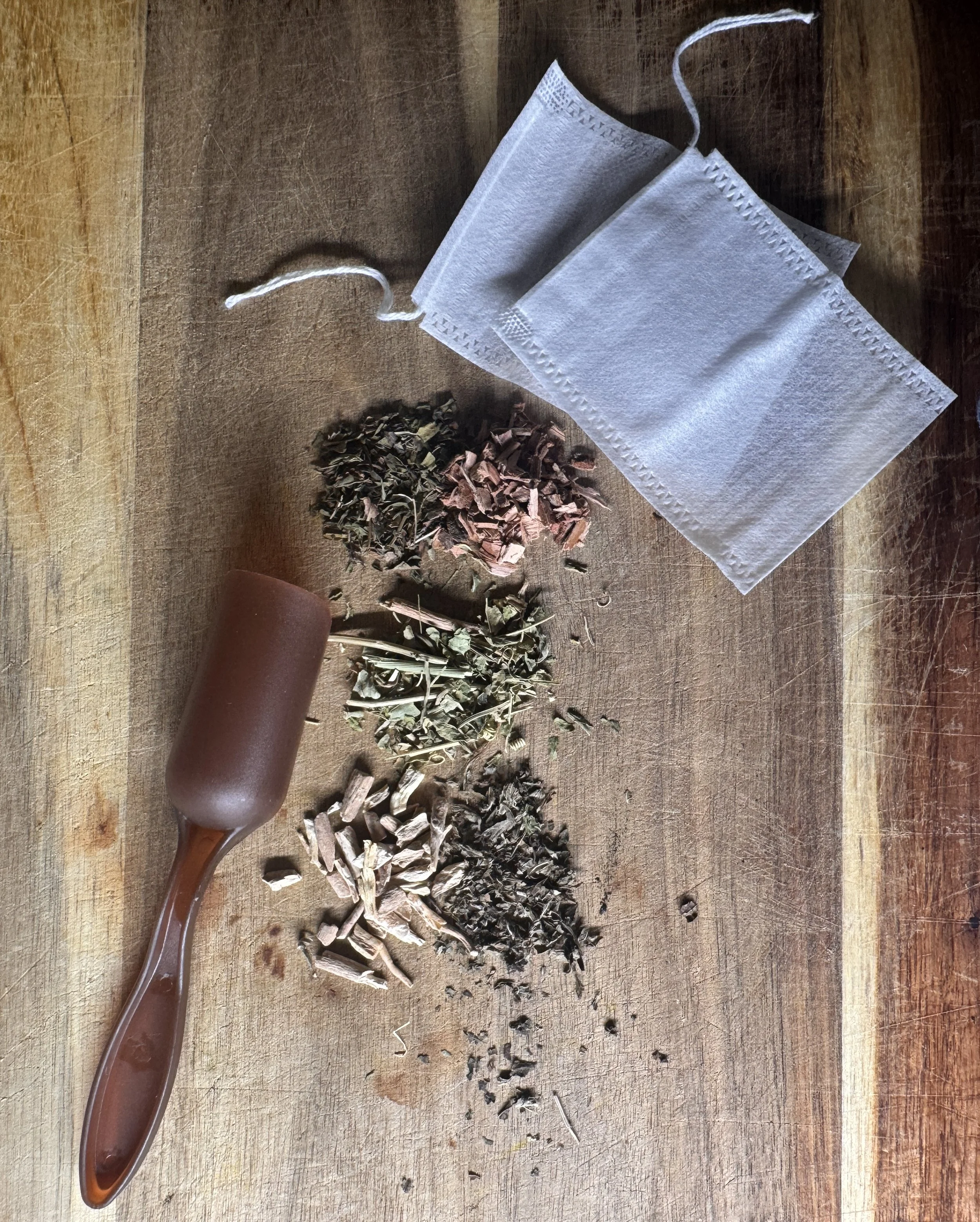 Three piles of dried herbs or tea leaves on a wooden surface, a brown spoon, and a white face mask.