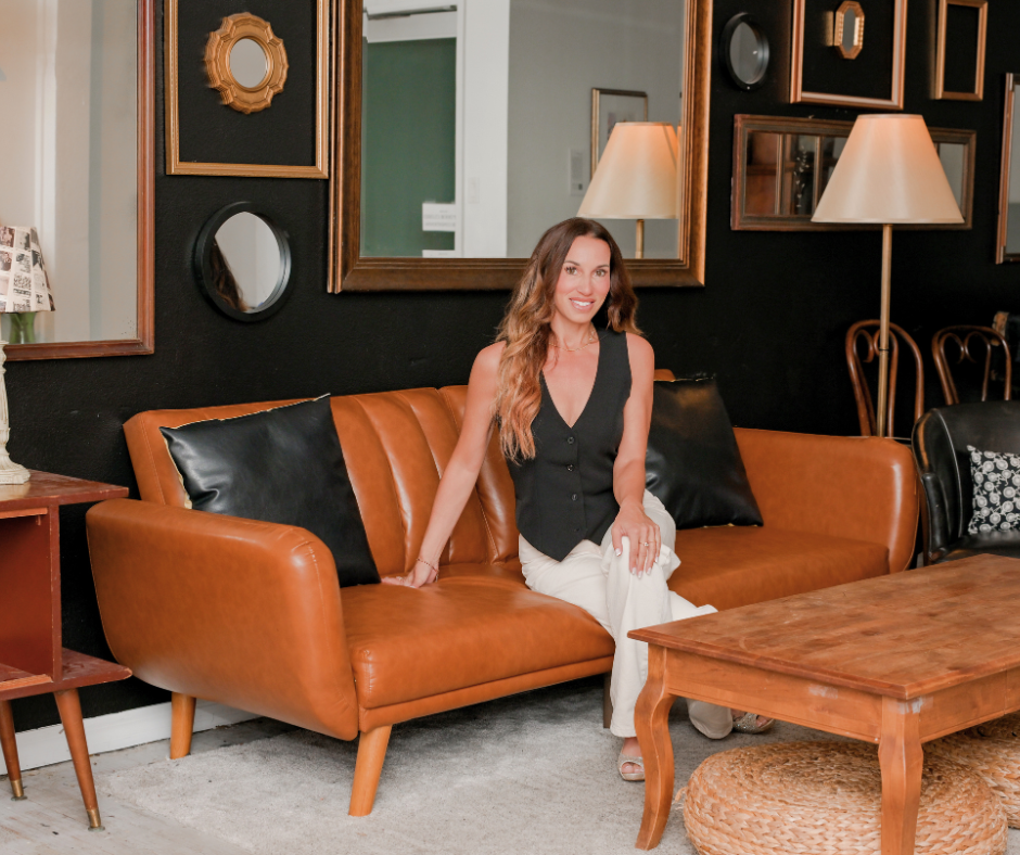 A woman sitting on a brown leather couch in a room with black walls decorated with mirrors and framed art, next to a wooden coffee table and two standing lamps.