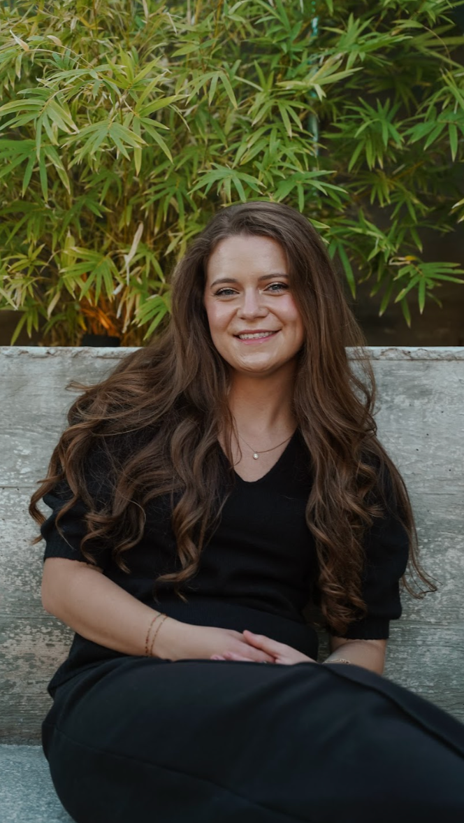 Professional portrait of a smiling woman with long brown hair wearing a black blouse and pants, seated on a wooden bench with green bamboo plants in the background.