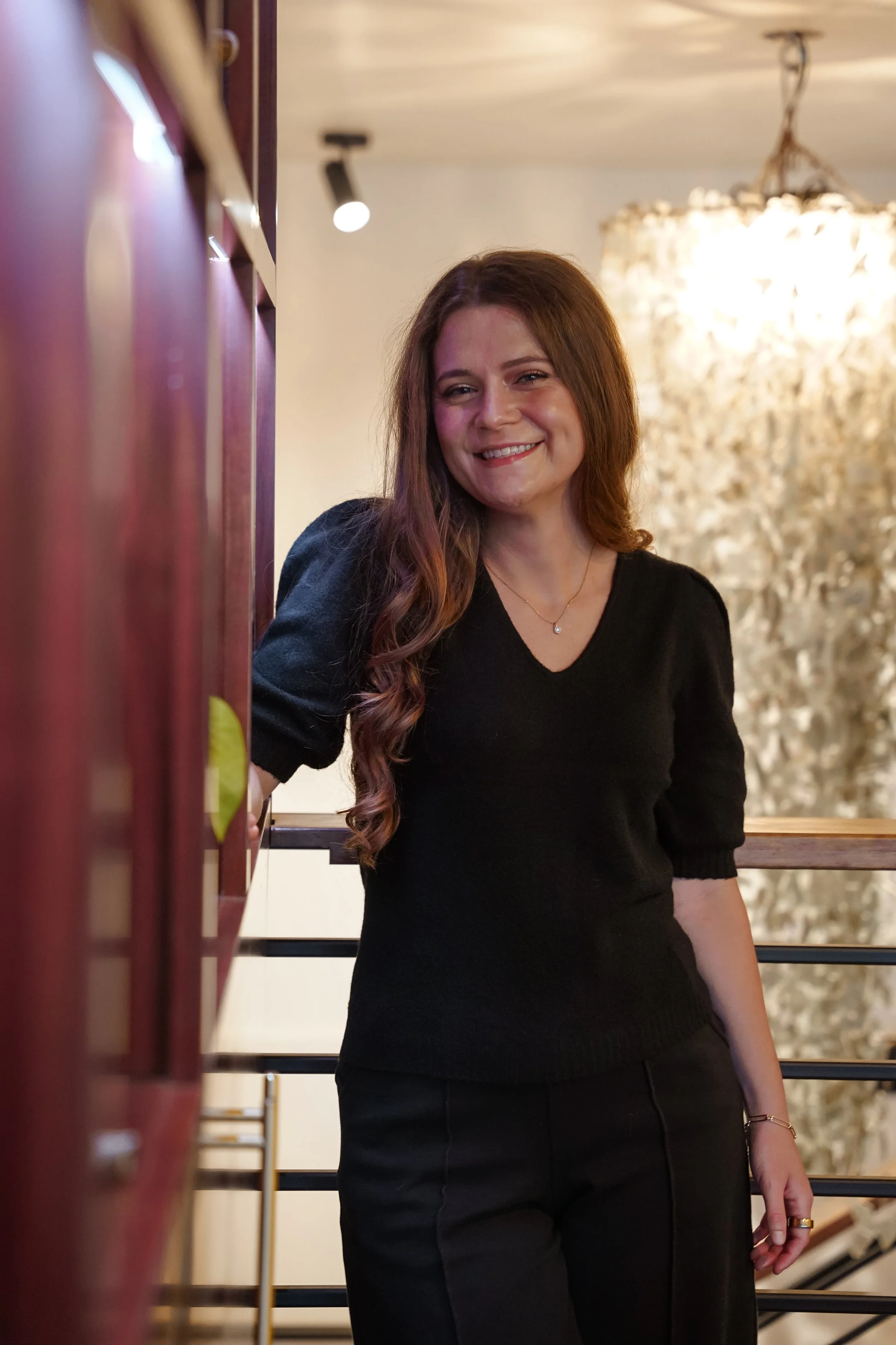 A woman with long, wavy red hair smiling and posing indoors, wearing a black top and black pants, with a chandelier and decorative wall behind her.