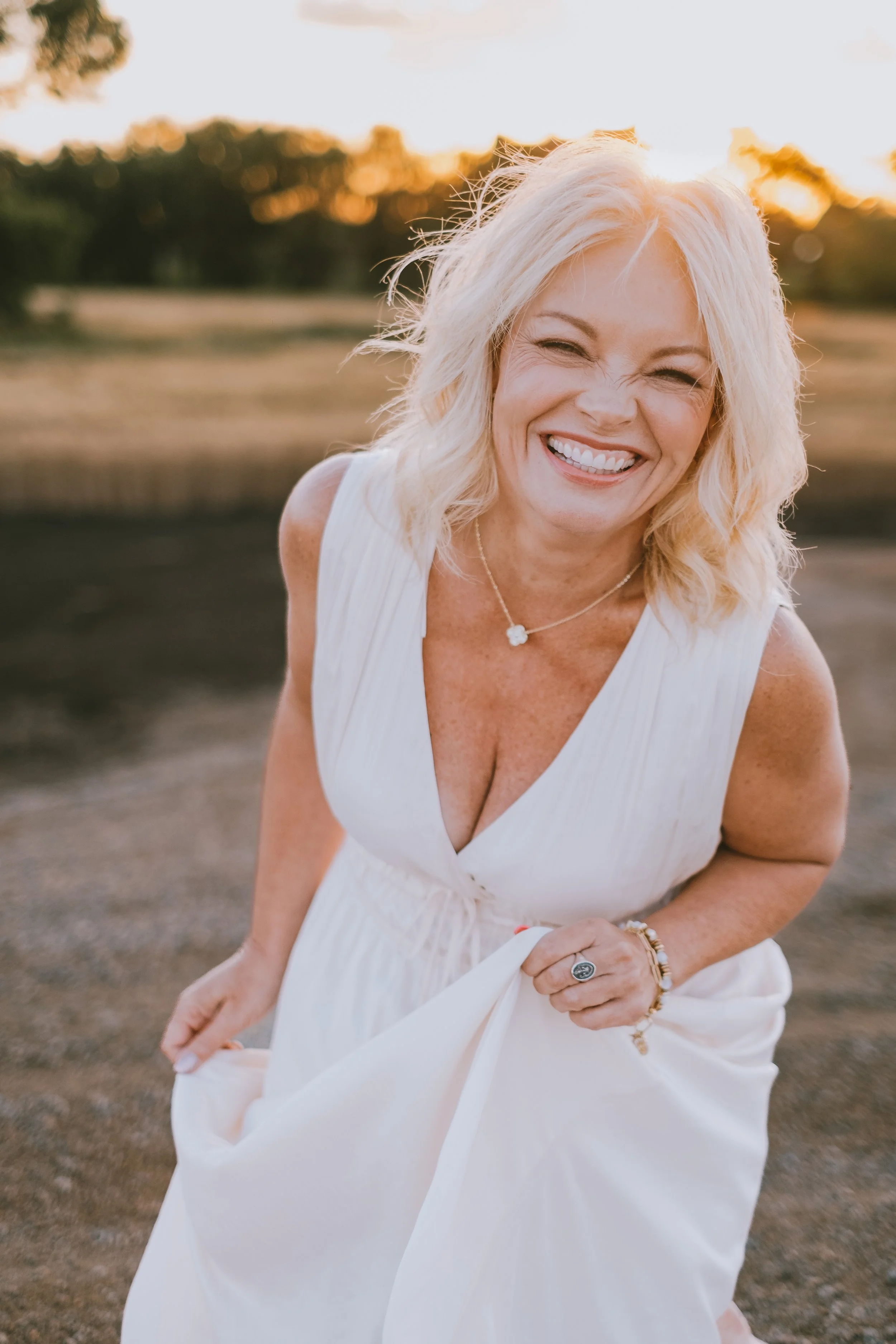 A woman with blonde hair wearing a white sleeveless dress, smiling and holding the dress with one hand outdoors during sunset, with trees and sky in the background.